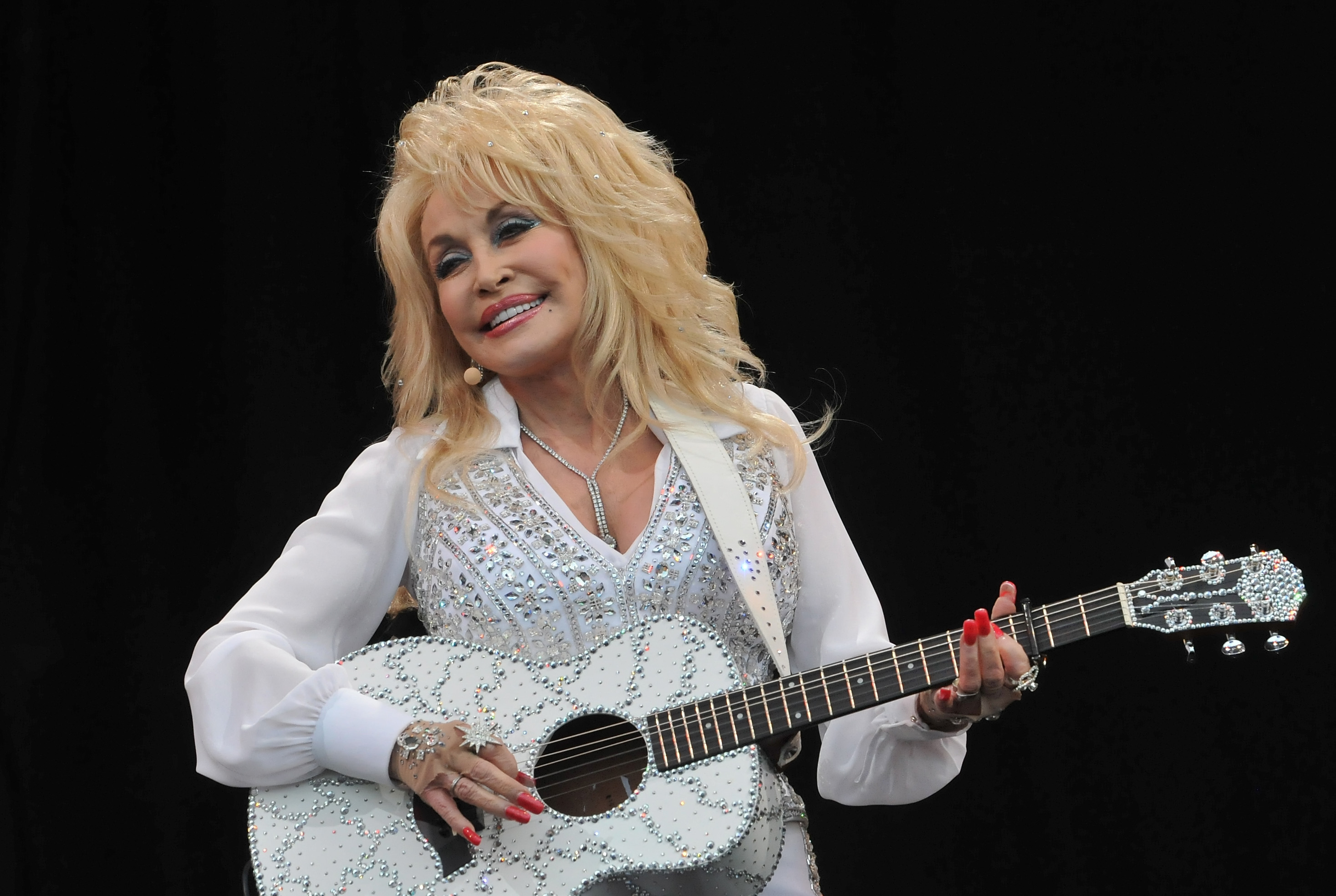 Dolly Parton wears a white rhinestoned shirt and holds a matching guitar.