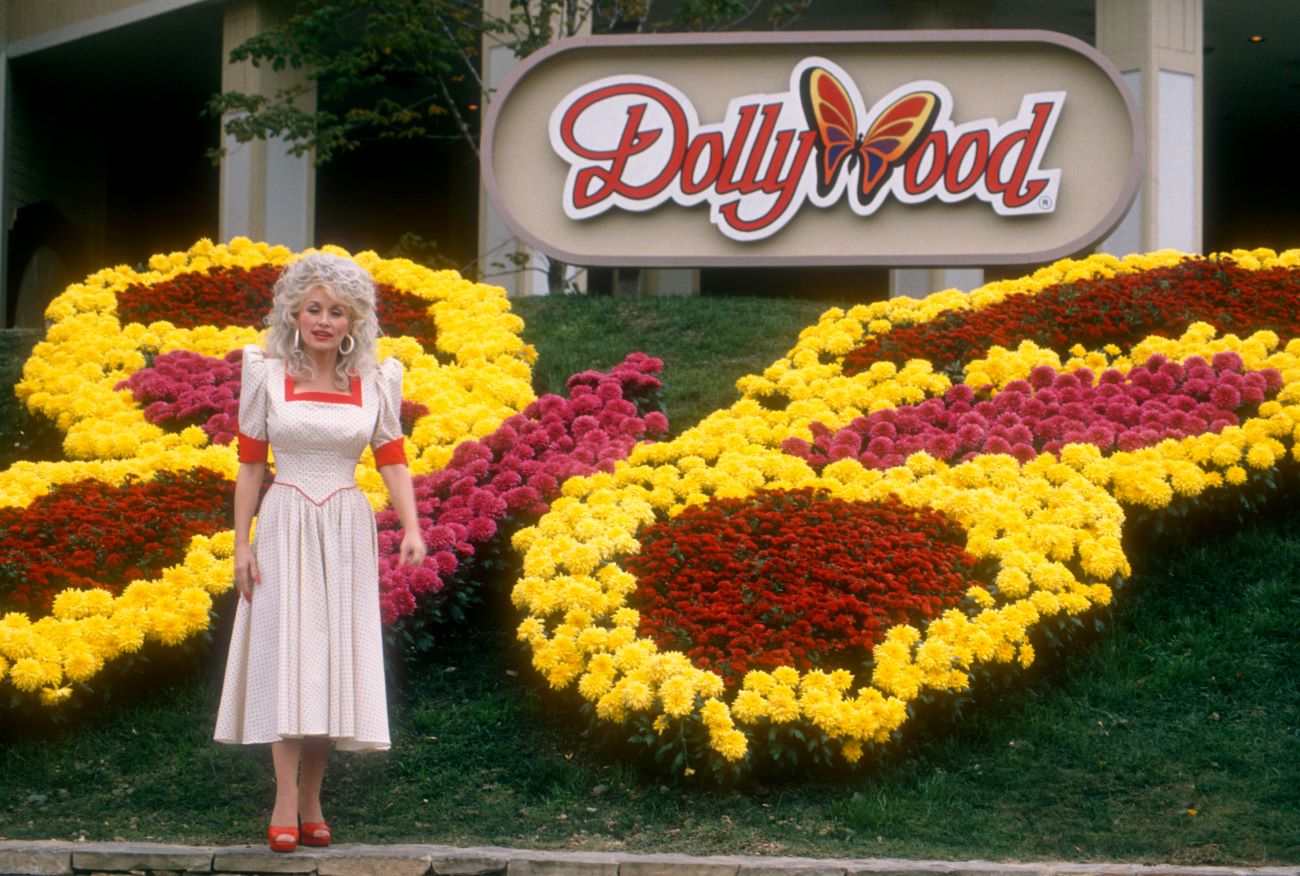 Dolly Parton stands in front of the Dollywood sign and flowers planted in the shape of a butterfly.