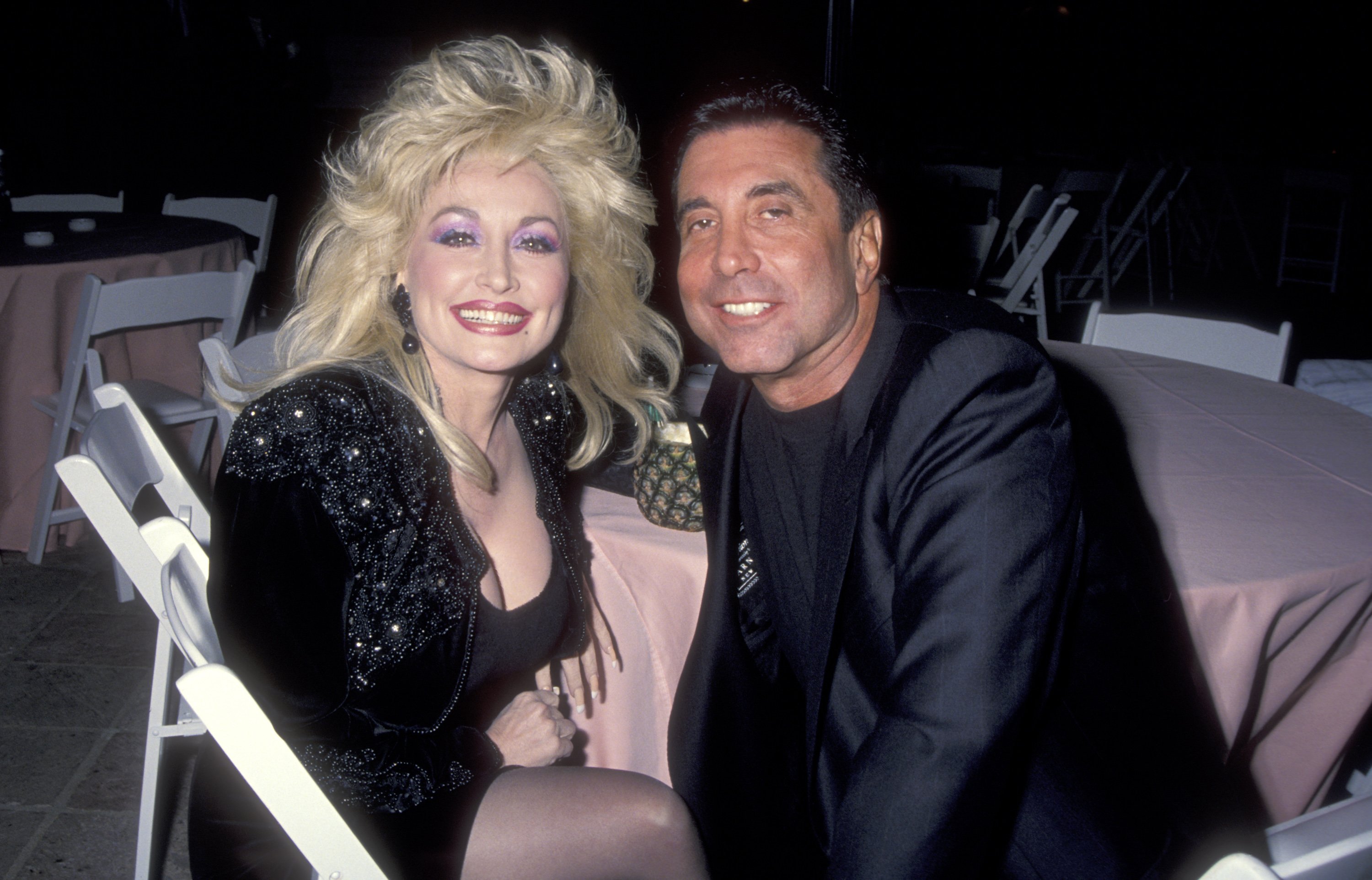 Dolly Parton and her manager Sandy Gallin wear black and sit at a round table with a pink tablecloth. 