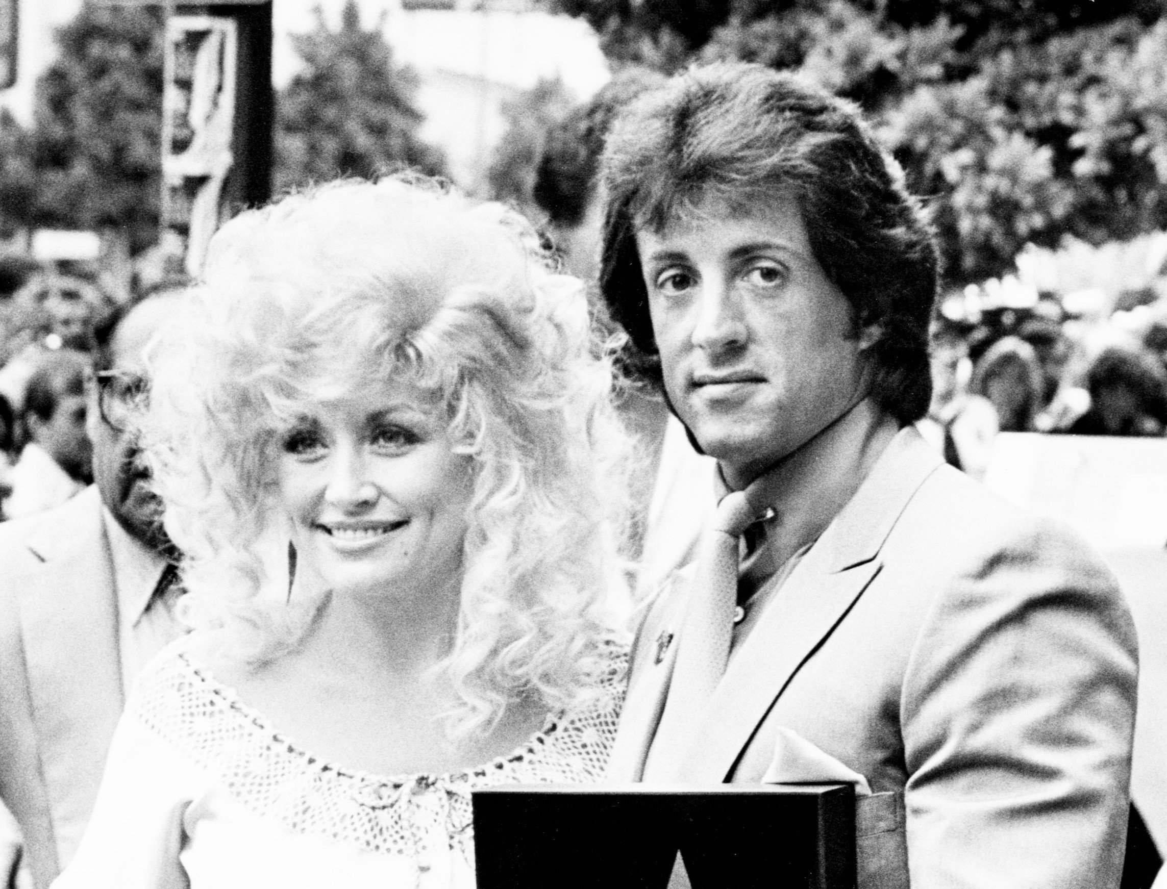 A black and white picture of Dolly Parton and Sylvester Stallone posing together. She wears a white dress and he wears a suit.
