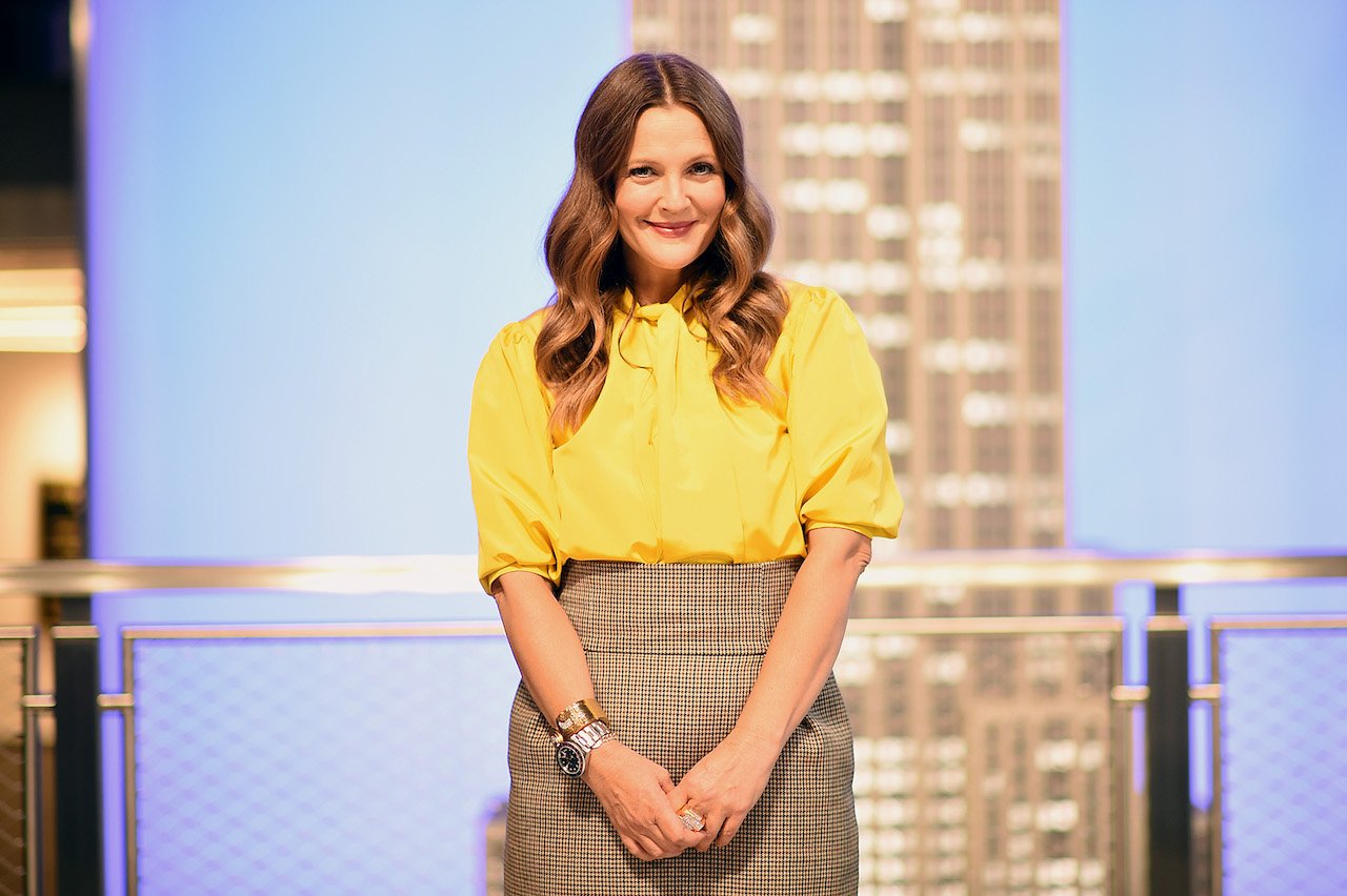 Drew Barrymore poses in a yellow top with her hands together in front of her 