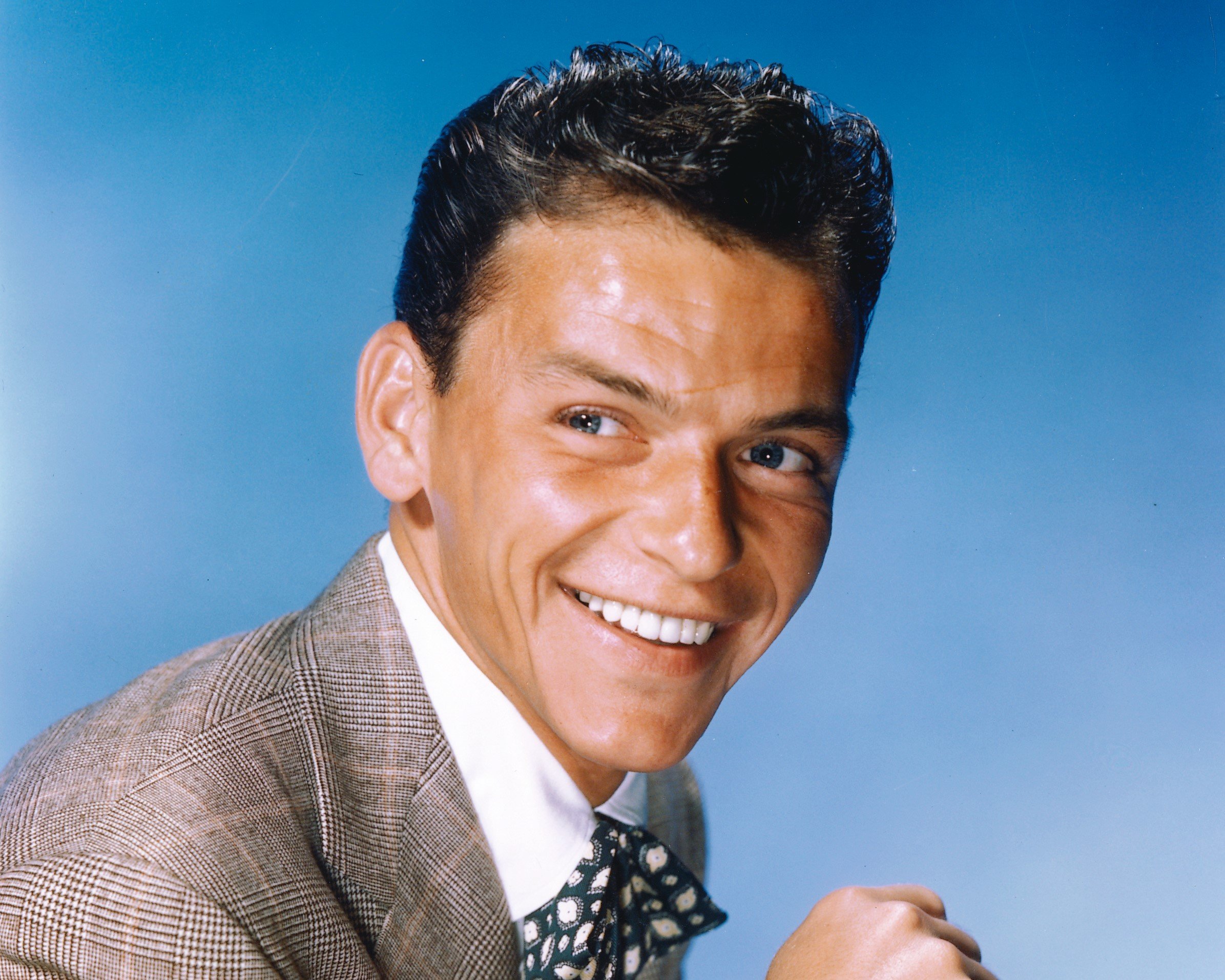 Frank Sinatra wears a suit and smiles against a blue background.