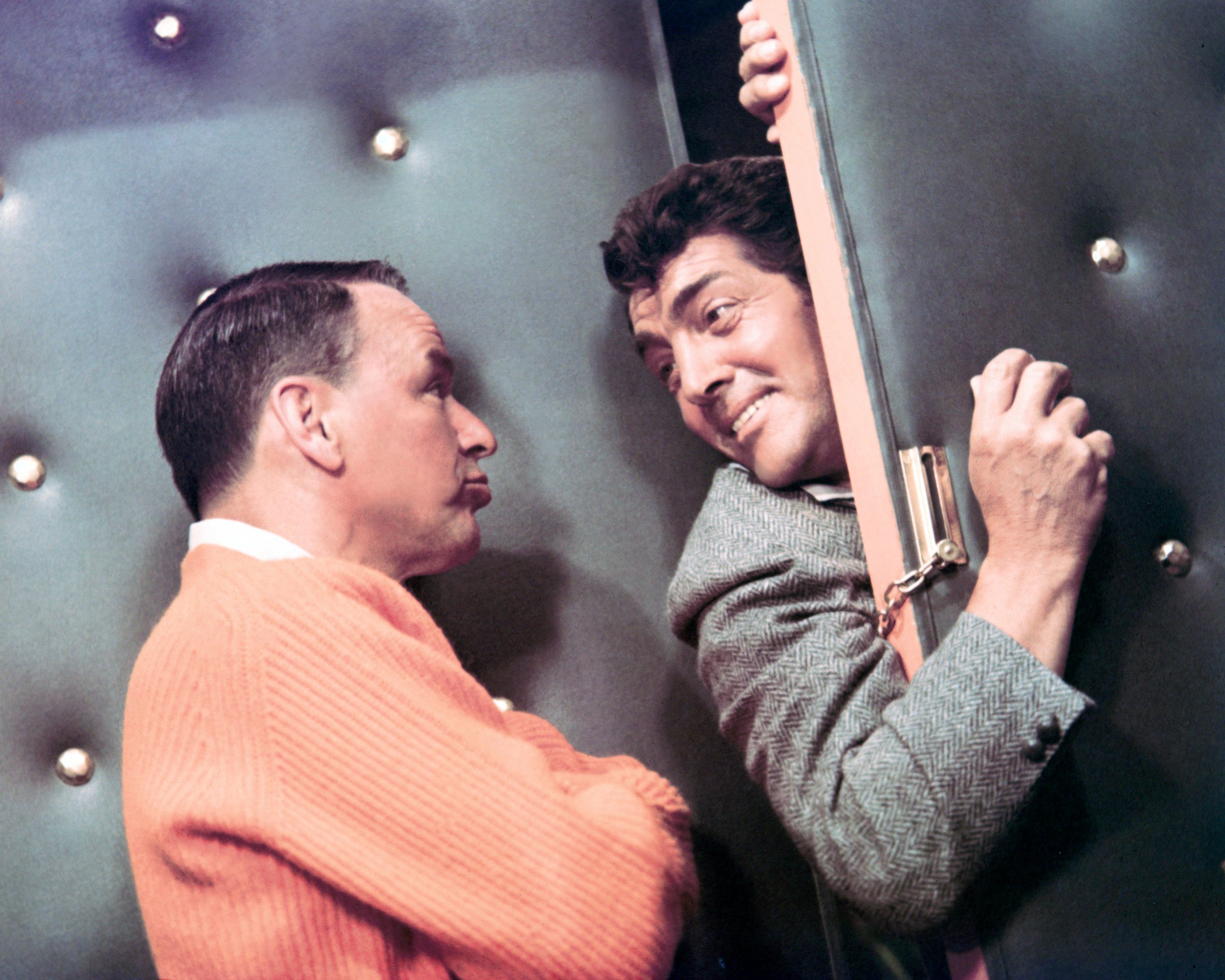 Dean Martin pokes his head and shoulder in through a locked door. Frank Sinatra stands with his arms folded and faces Martin.