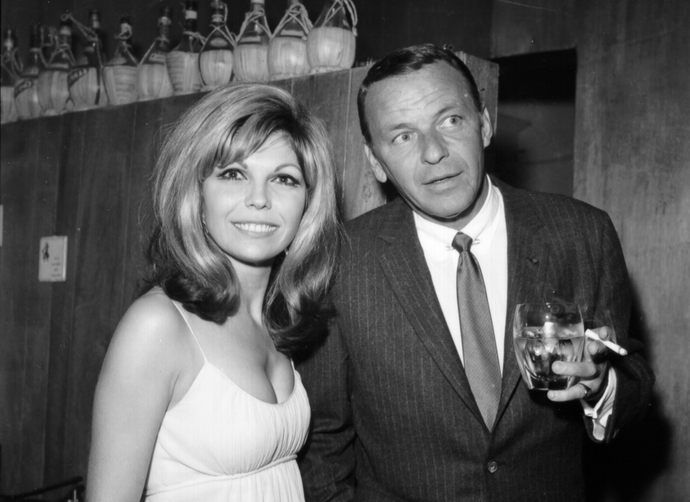 A black and white photo of Nancy Sinatra and Frank Sinatra. He holds a drink and a cigarette.