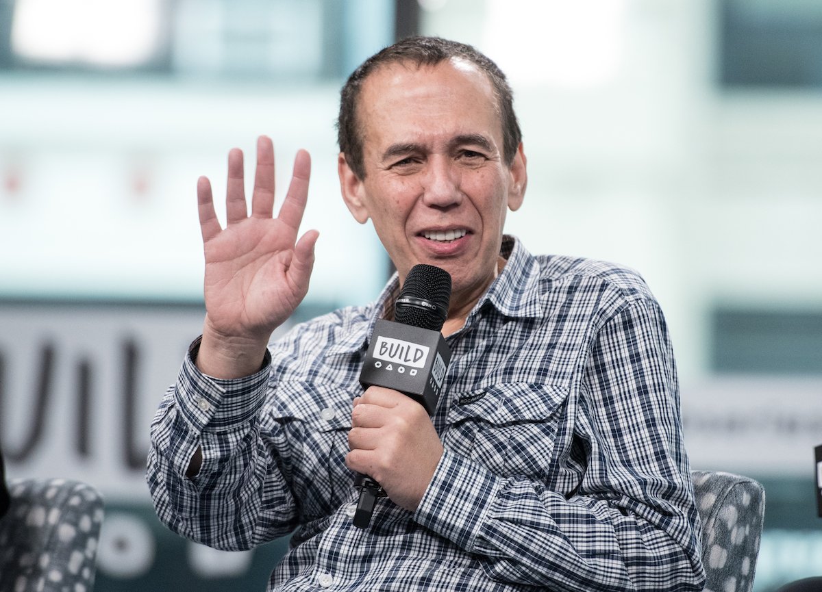 Gilbert Gottfried waves to the camera and holds a microphone.
