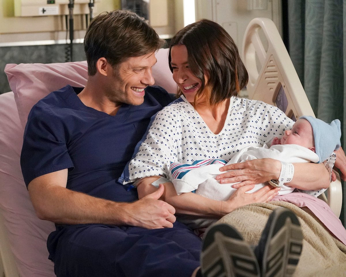 Chris Carmack and Caterina Scorsone smiling, sitting in a hospital bed, holding a baby
