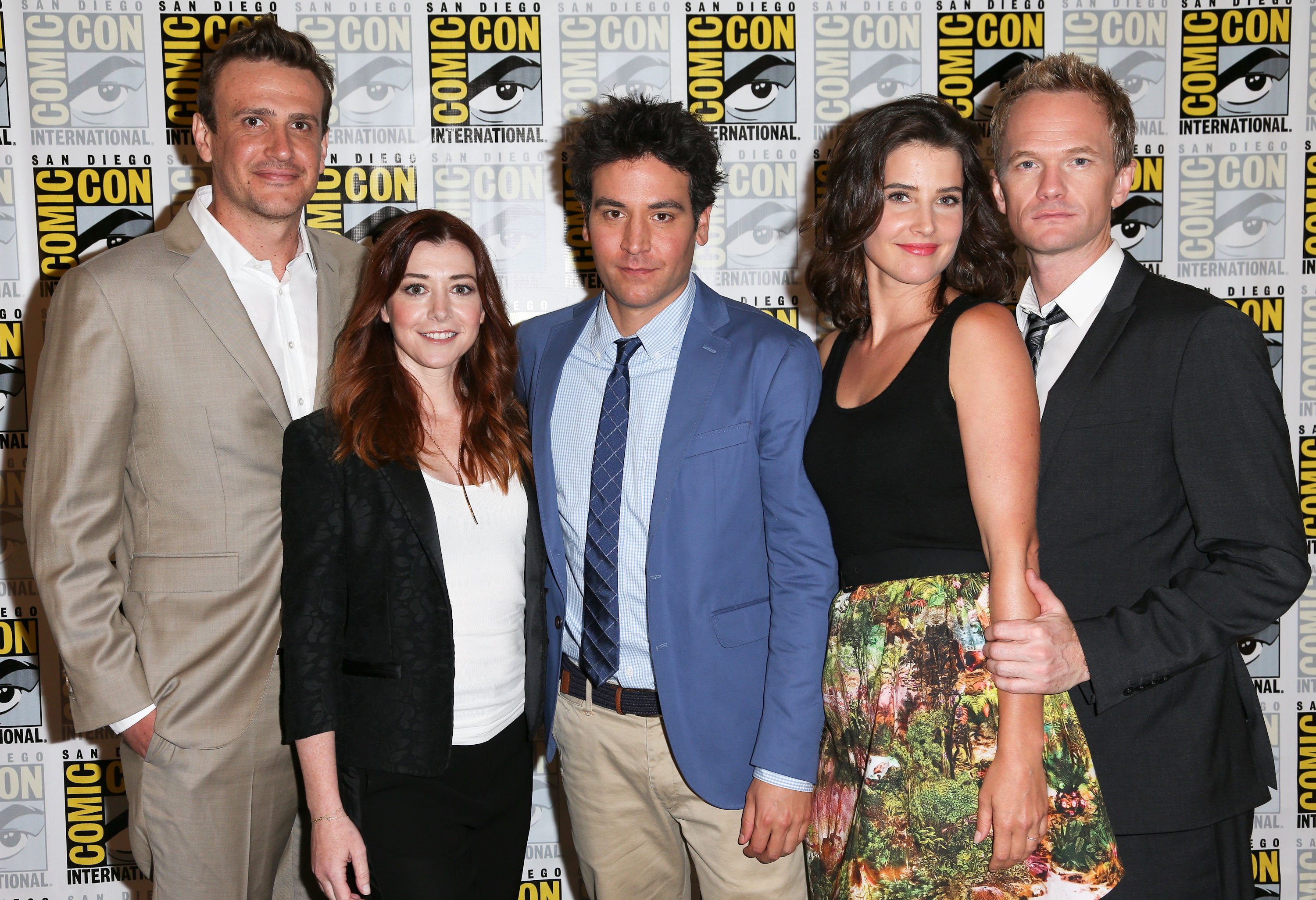 The cast of 'How I Met Your Mother,' Jason Segel, Alyson Hannigan, Josh Radnor, Cobie Smulders, and Neil Patrick Harris, who might appear in 'How I Met Your Father' Season 2, pose for photos. Segel wears a tan suit over a white button-up shirt and tan pants. Hannigan wears a black jacket over a white shirt and black pants. Radnor wears a blue suit over a light blue button-up shirt, blue tie, and tan pants. Smulders wears a black tank top and skirt with a forest scene on it. Harris wears a black suit over a white button-up shirt and black and gray tie.