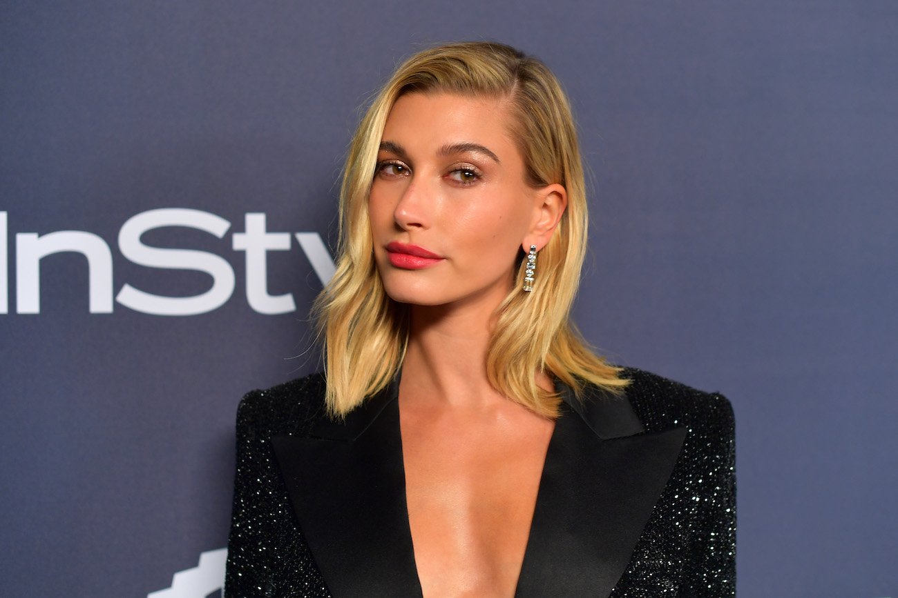 Hailey Bieber wearing a black outfit with plunging neckline and standing in front of a gray background