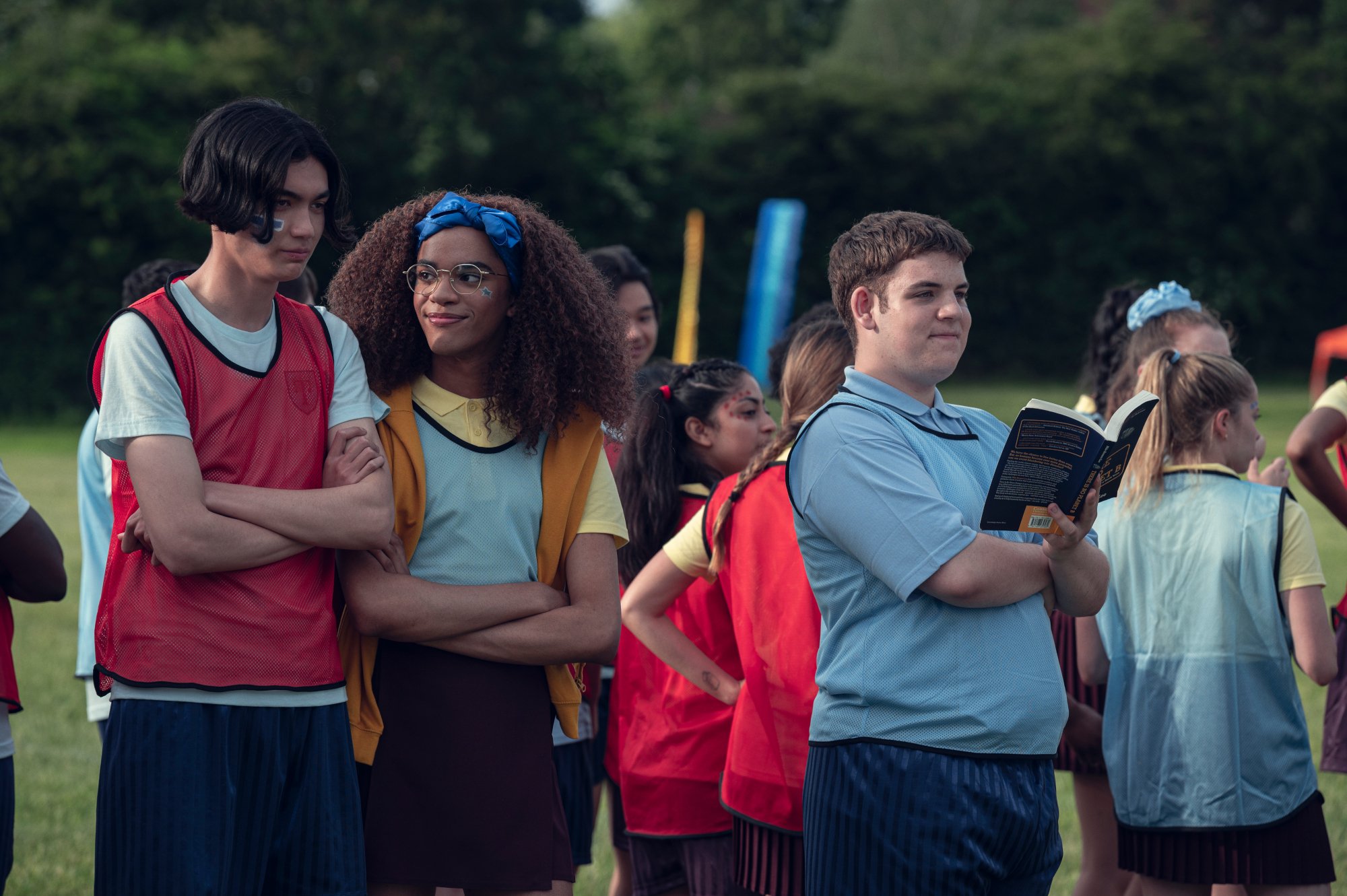 William Gao, Yasmin Finney, and Tobie Donovan as Tao, Elle, and Isaac in Netflix's TV show adaptation of the 'Heartstopper' books. They're standing next to one another outside, and there's a crowd of people behind them.