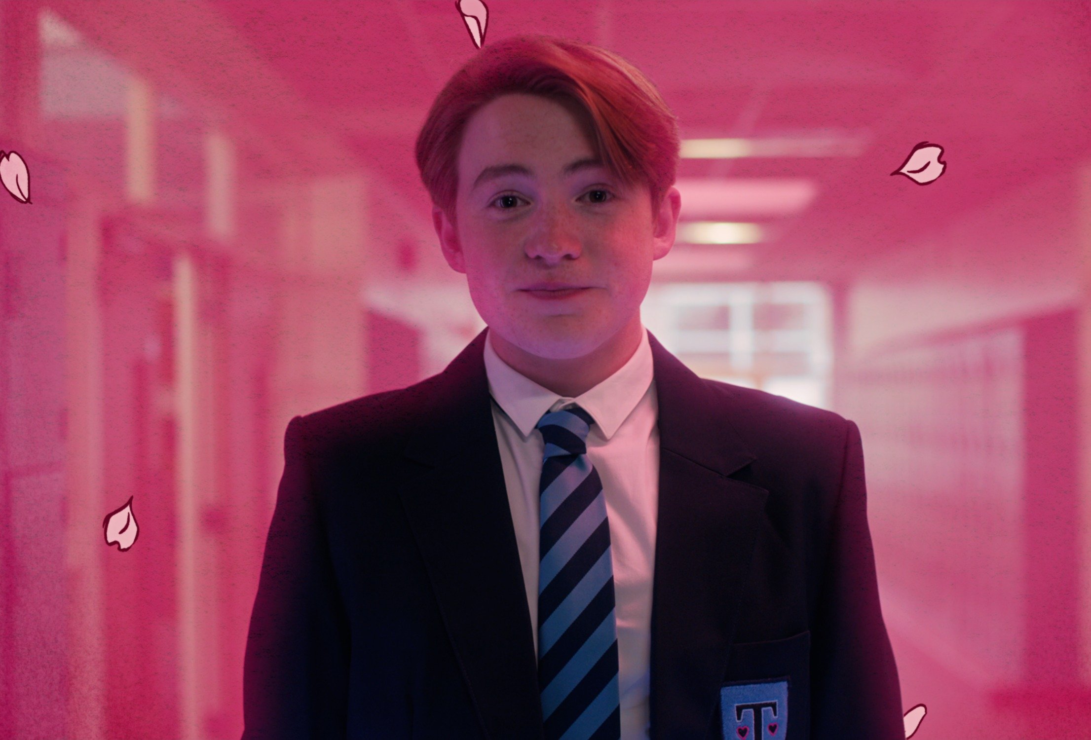 Kit Connor as Nick Nelson in Netflix's 'Heartstopper' show, which adapts Alice Oseman's books. He's wearing a school uniform and smiling, and everything around him has a pink lighting.