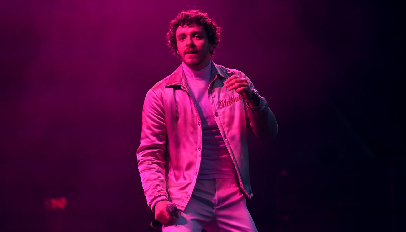 Jack Harlow is lit by pink light and stands, holding a microphone.
