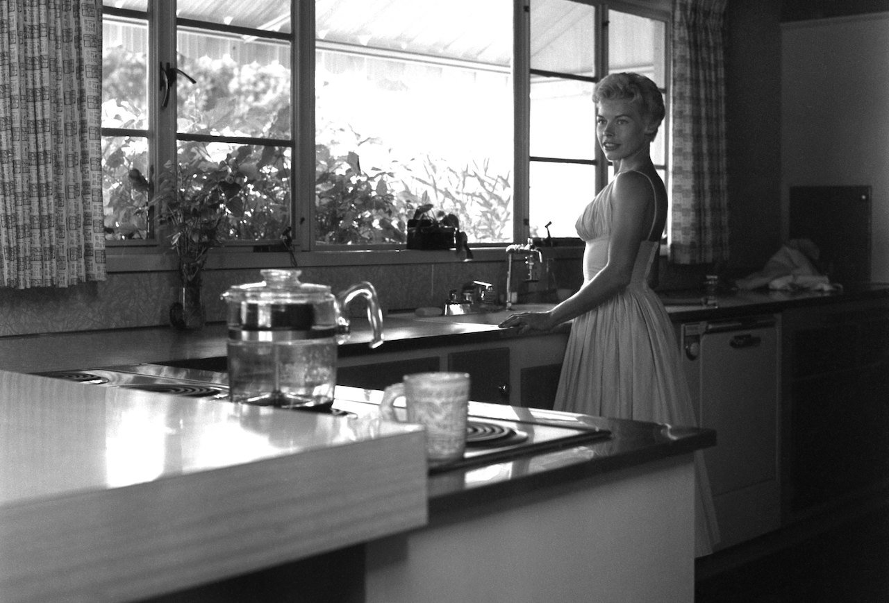 Jody Carson stands at the sink in her kitchen, in a dress, and looking over her shoulder c. 1956