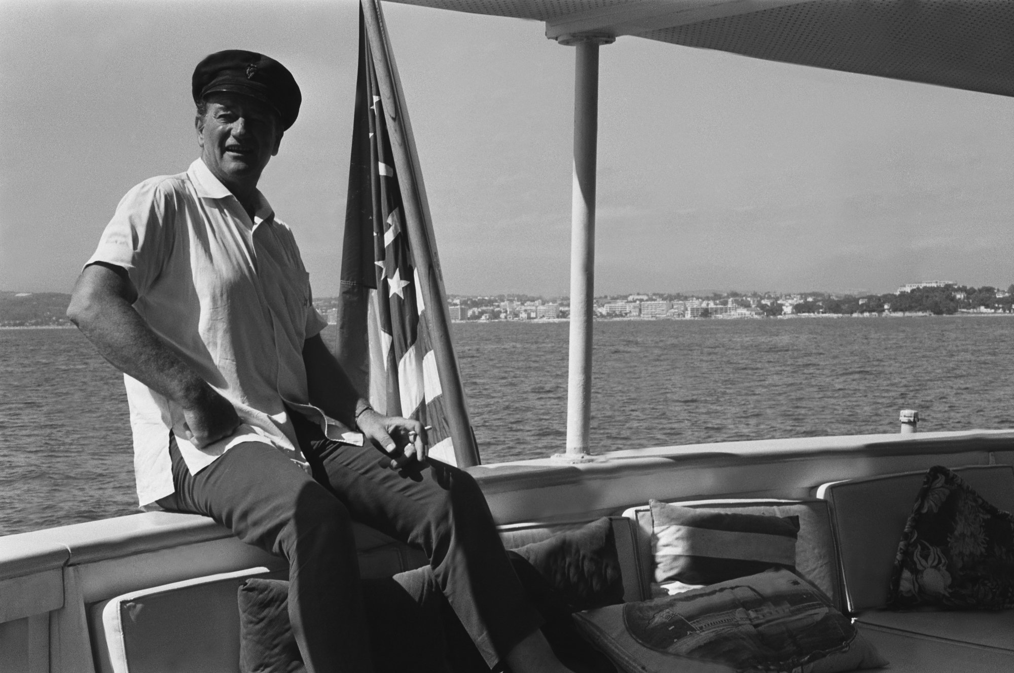 John Wayne aboard Wild Goose, his houseboat, wearing a collared shirt, captain hat, sitting next to American flag