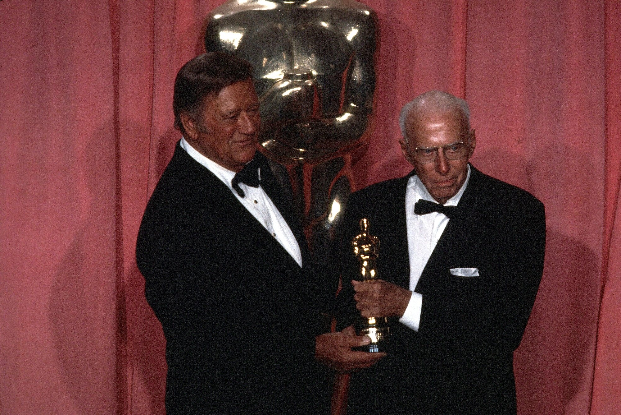 John Wayne and Howard Hawks at the 1975 Oscars holding the Honorary Award together in front of red curtains