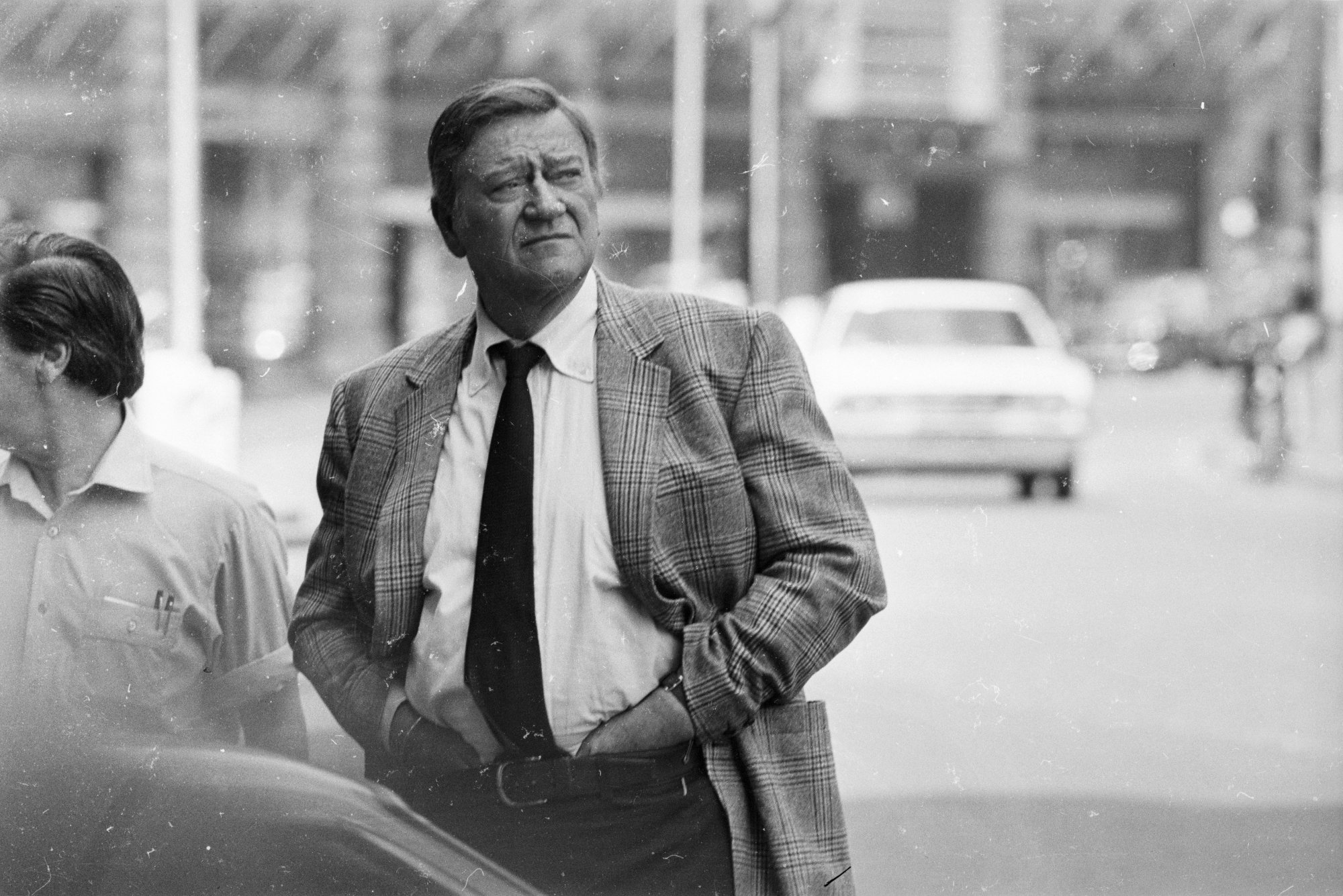 John Wayne standing with his height over six feet wearing a suit and tie with a car driving away in the background