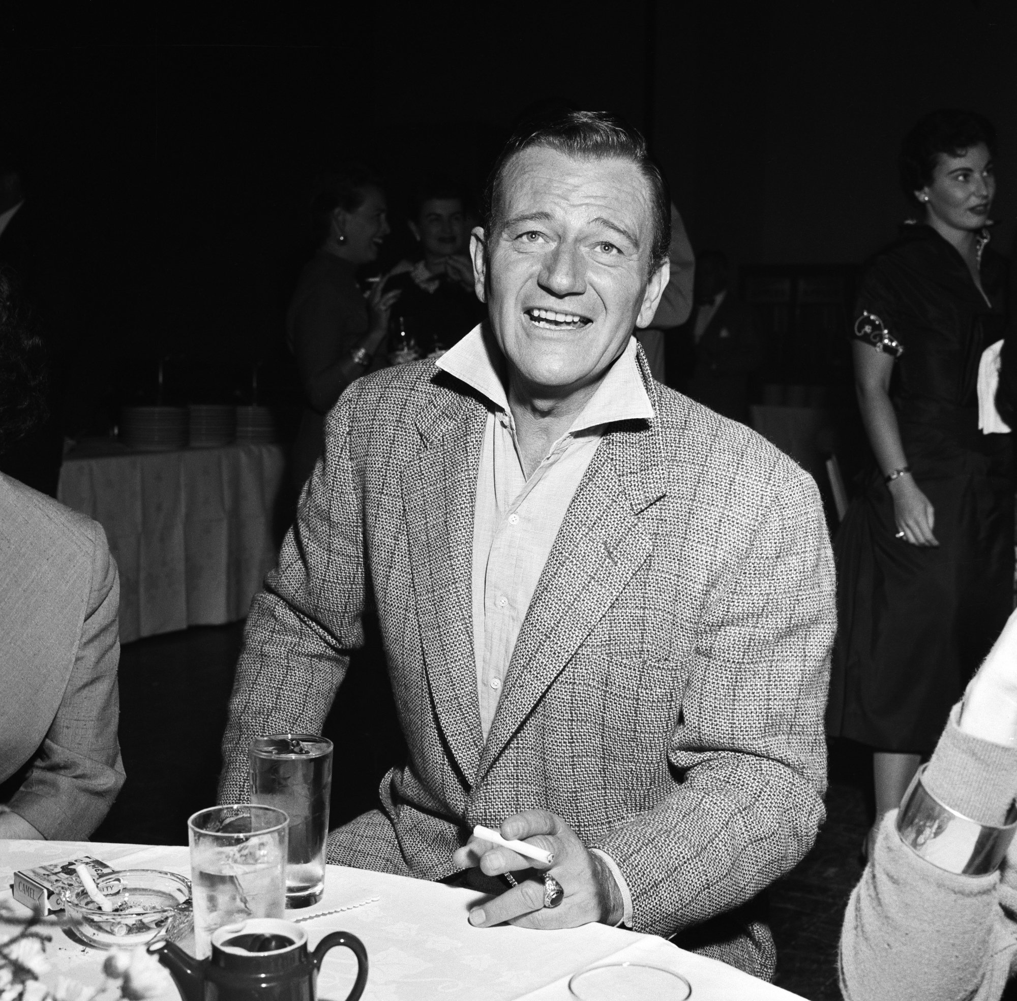 John Wayne, the actor who gave advice to Michael Caine, wearing a suit with a cigarette in his hand, sitting at a table