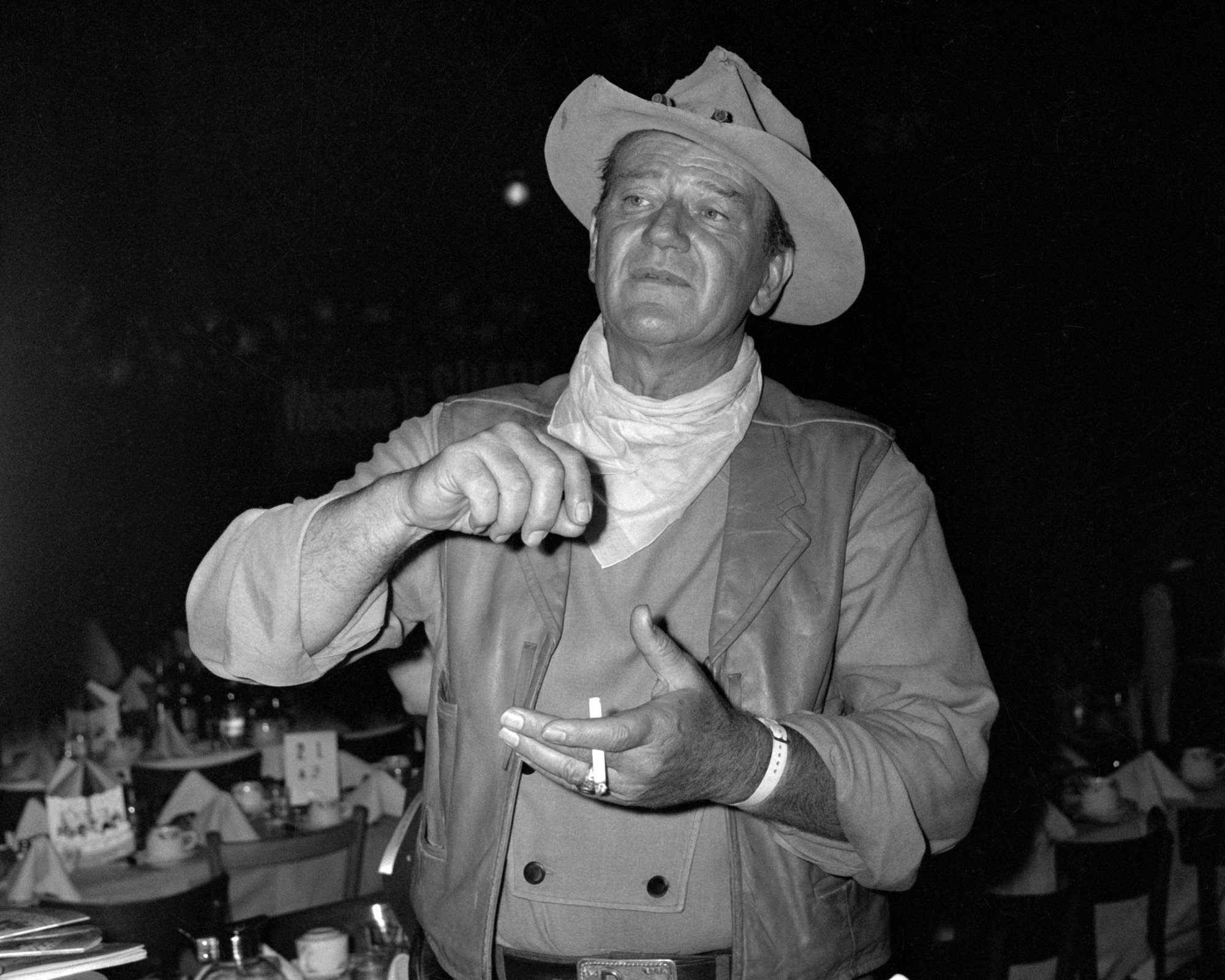 John Wayne, who submitted the John Wayne Casserole recipe, wearing a cowboy hat and holding a cigarette at an unknown dinner event