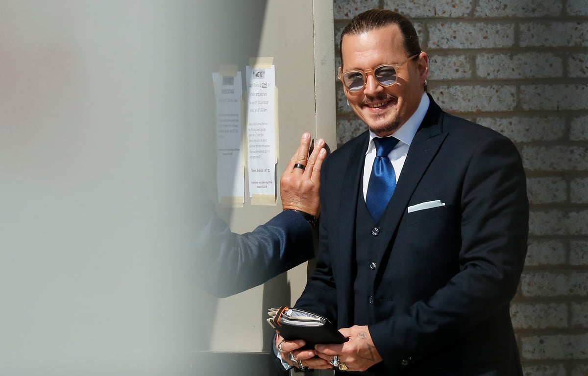 Johnny Depp stands outside of the courtroom in a suit while smiling.