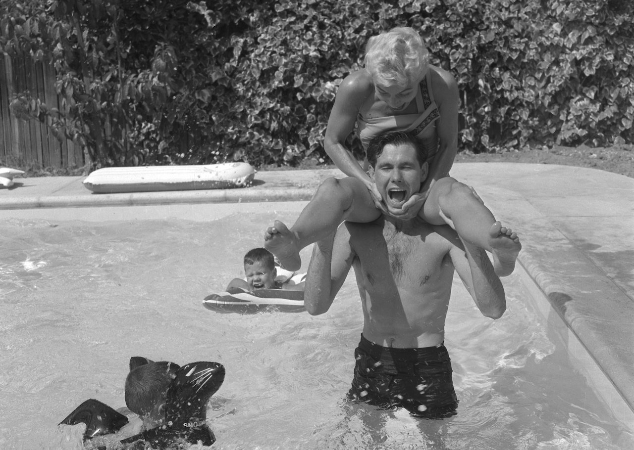 Johnny Carson with his wife, Jody, sitting on his shoulders, playing in their pool with their sons c. 1956