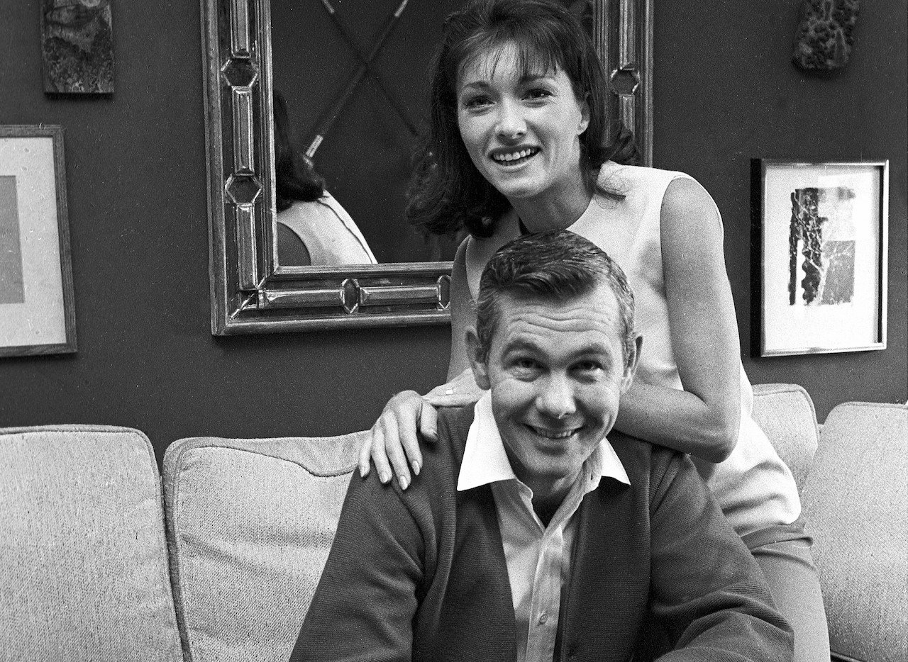 Joanne Carson leans on Johnny Carson's back as the couple smiles and poses for a picture on their couch c. 1965