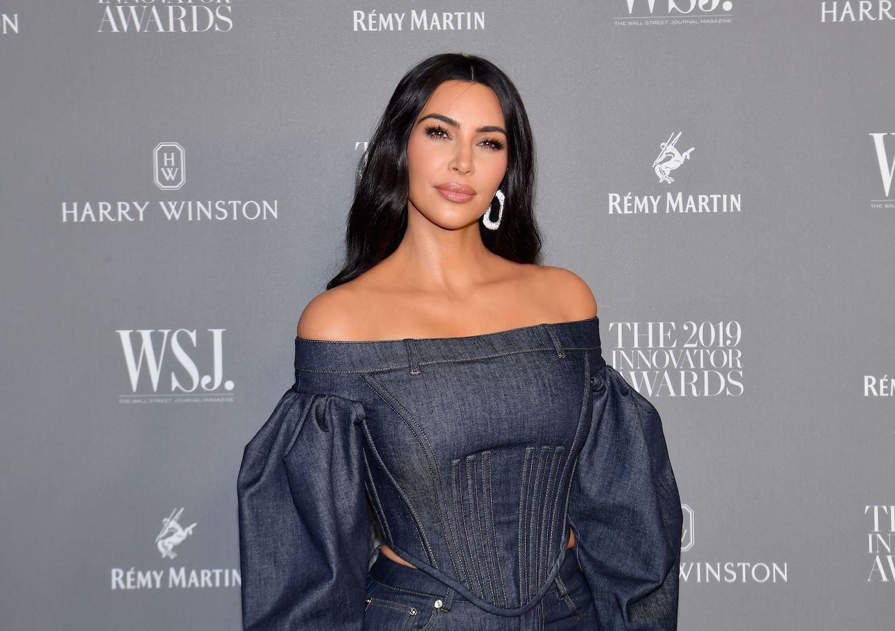 Kim Kardashian wearing an off-shoulder black outfit and standing in front of a gray background