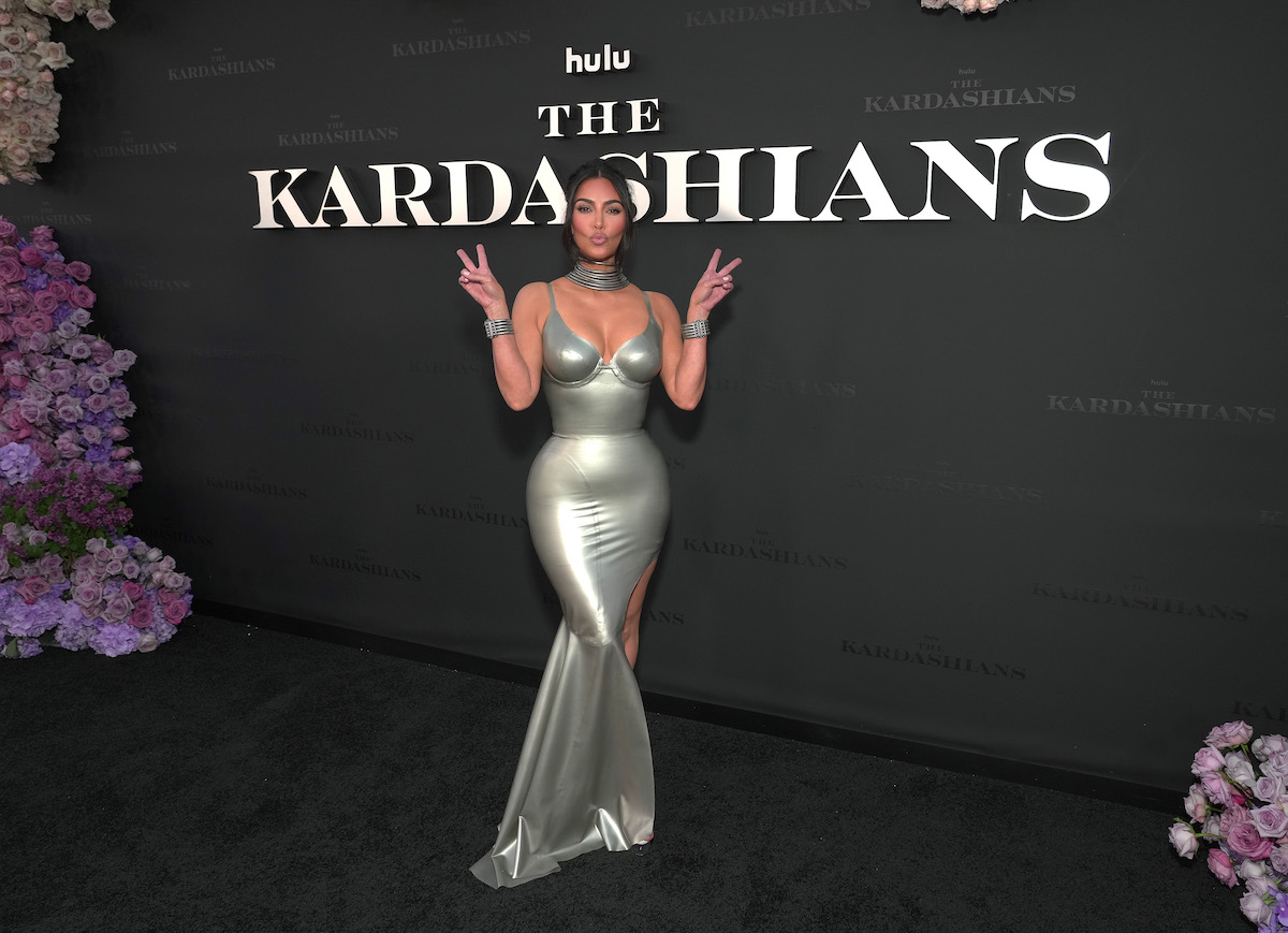 Kim Kardashian poses making a kissing face and throwing up two peace signs in front of a wall that says "Hulu The Kardashians."