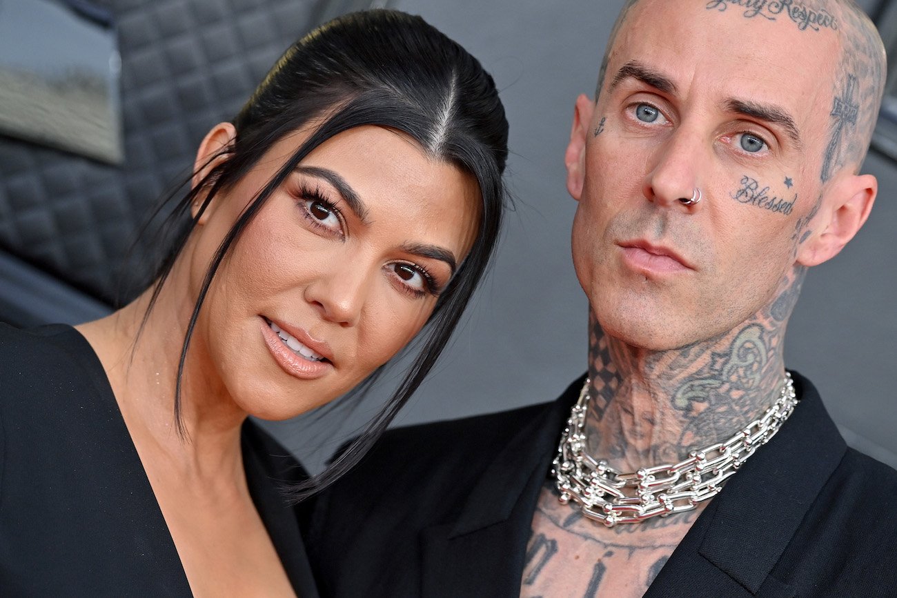 Close up shot of Kourtney Kardashian and Travis Barker. Both wearing black and standing in front of a gray background