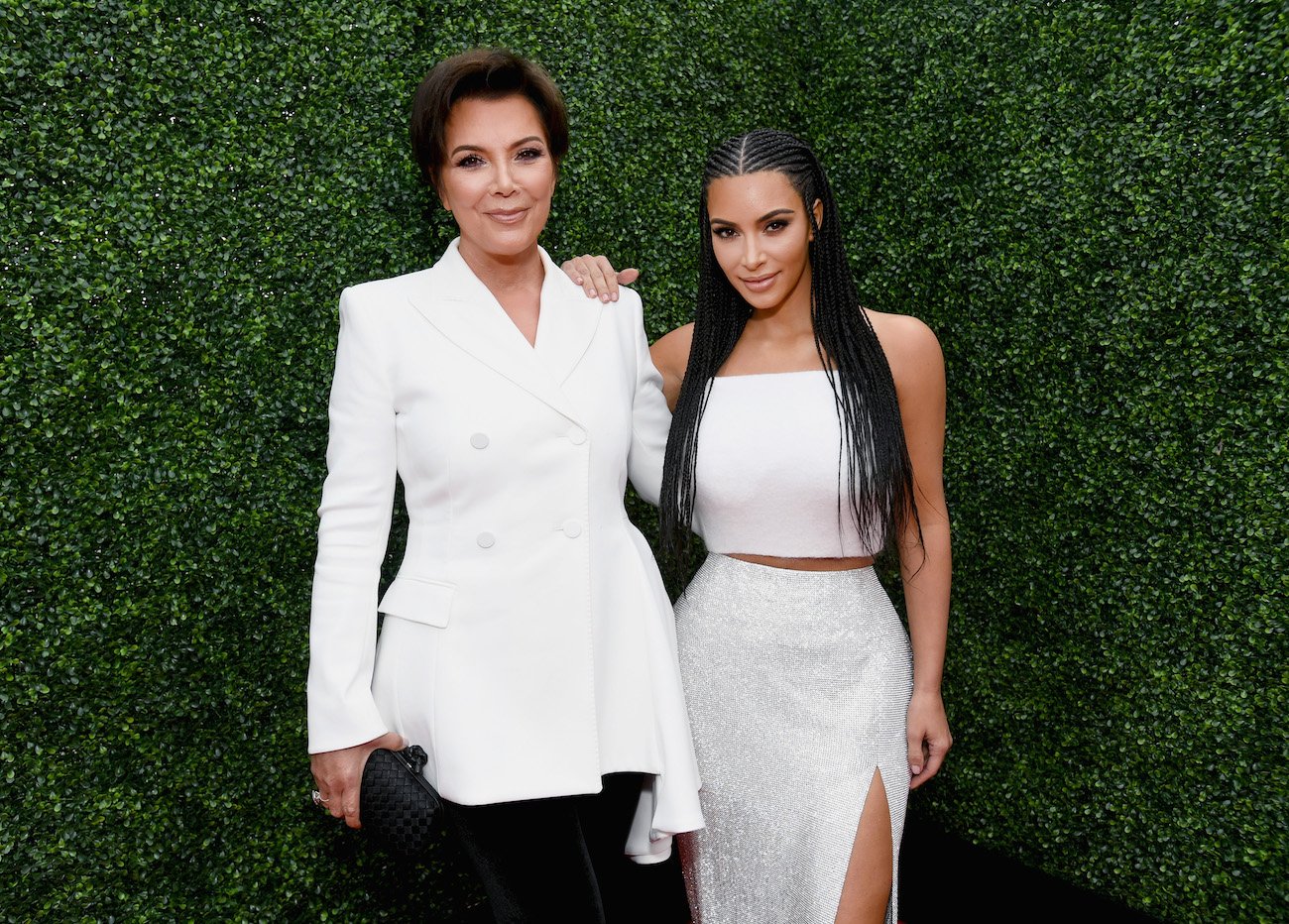 Kris Jenner and Kim Kardashian standing next to each other in white outfits and in front of a dark green background