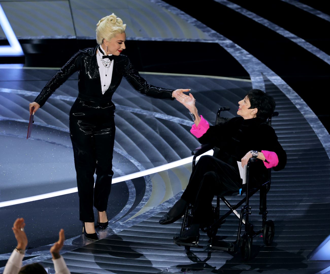 Lady Gaga wears a sparkling tuxedo and touches Liza Minnelli's hand on the Oscar's stage. She wears a black outfit with pink sleeves.