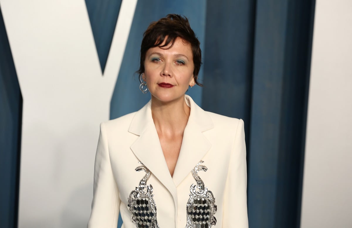 Maggie Gyllenhaal smirking while wearing a white dress.