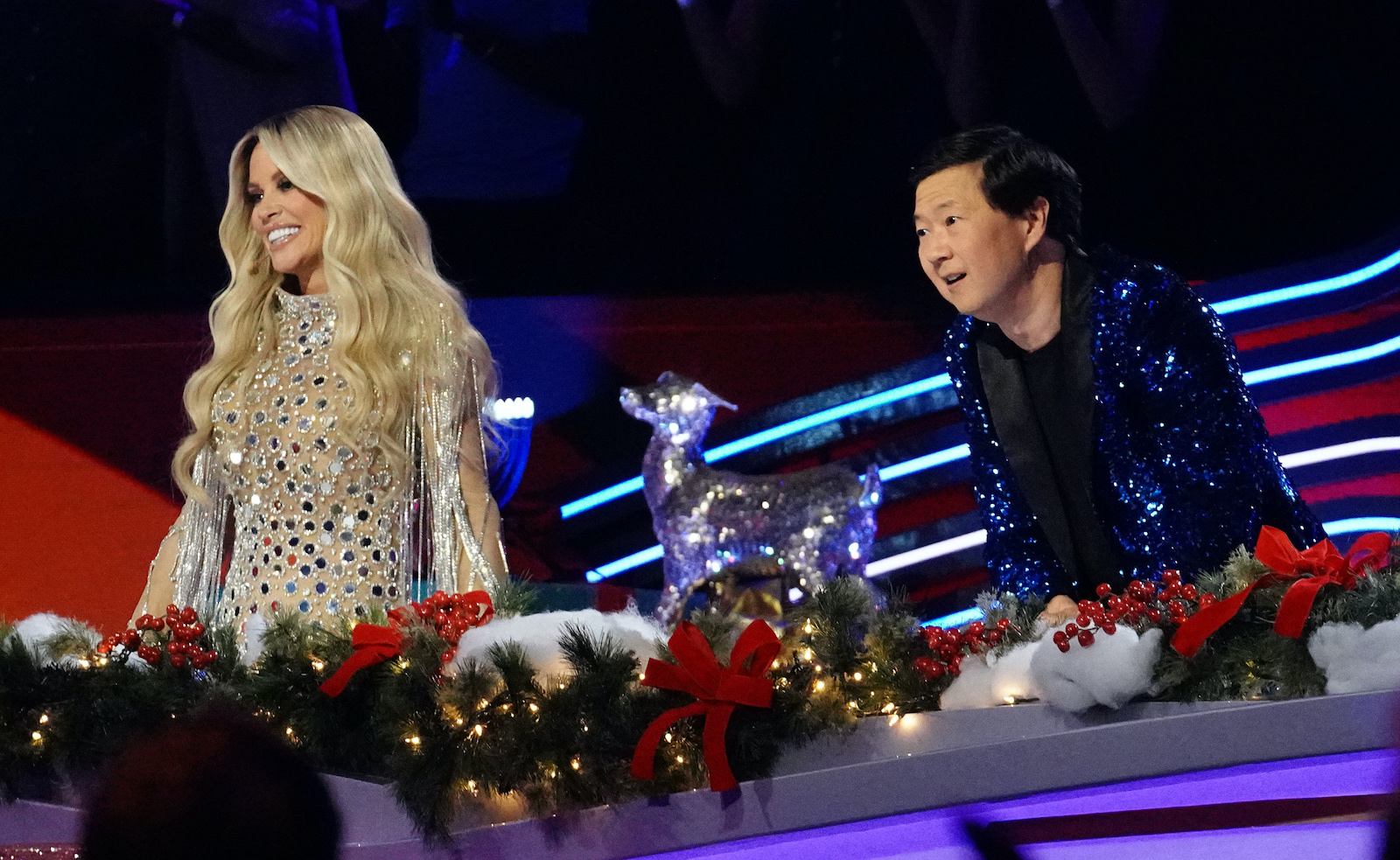Jenny McCarthy and Ken Jeong smile behind 'The Masked Singer' judge table.
