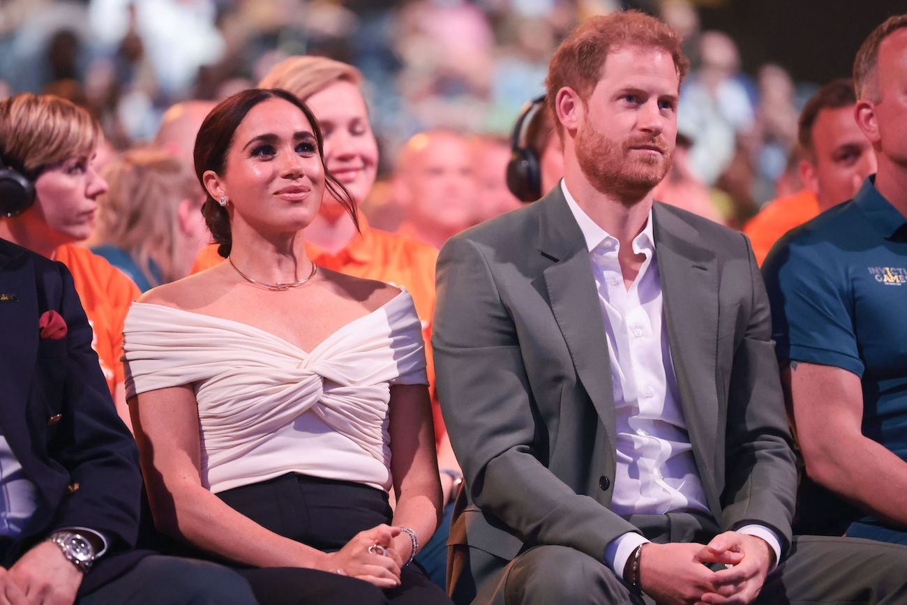 Meghan Markle wearing a white top and sitting next to Prince Harry, who is wearing a gray suit