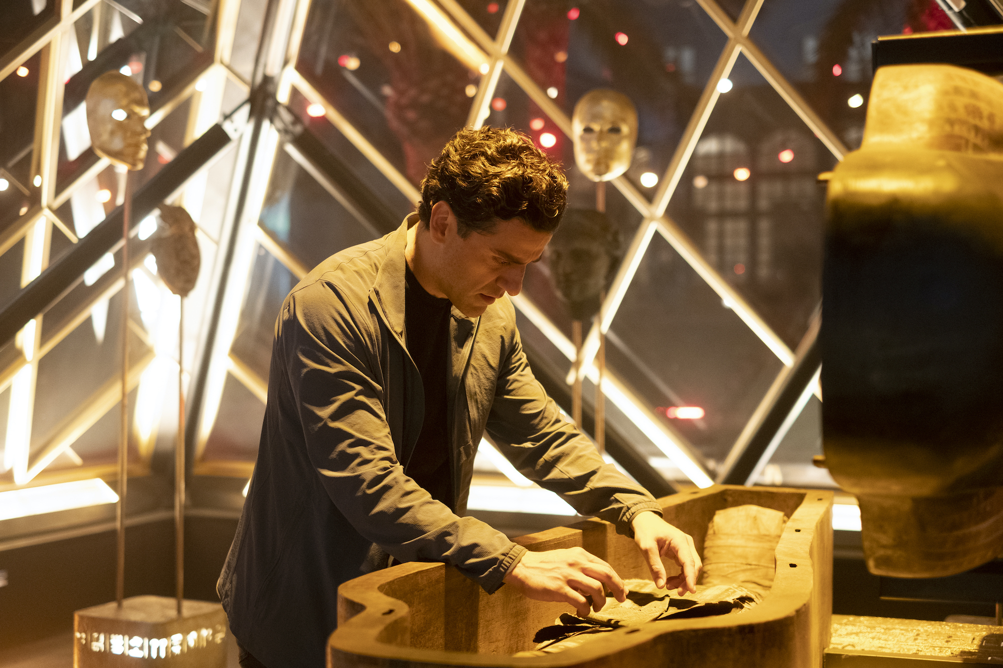 Oscar Isaac, in character as Marc Spector in the Easter egg filled 'Moon Knight,' wears a gray jacket over a black shirt while searching through a sarcophagus.