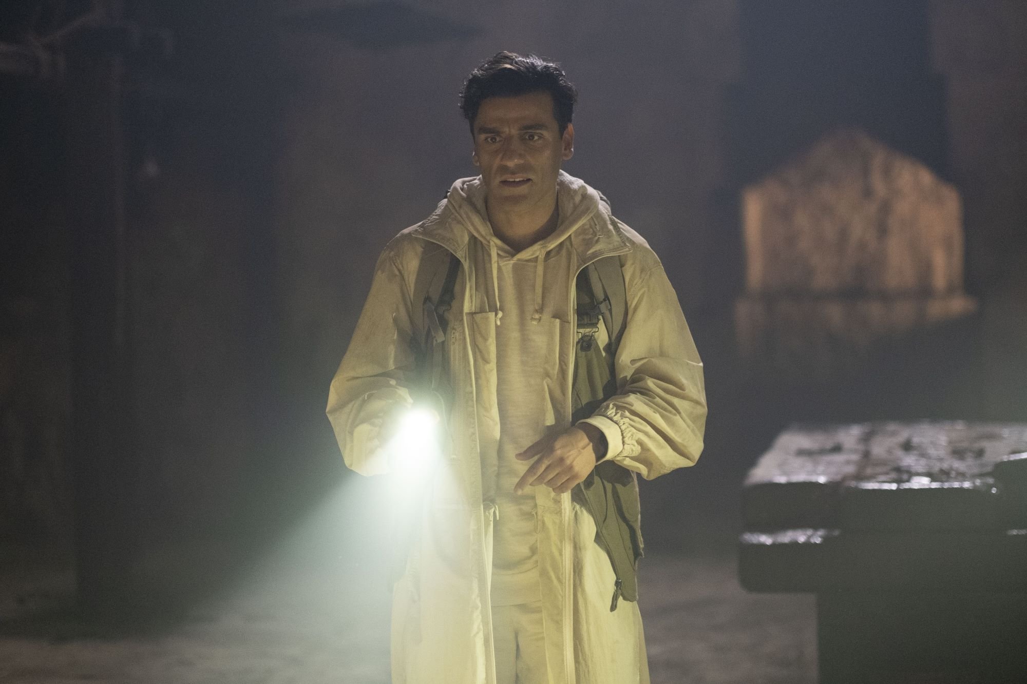 Oscar Isaac stars as Marc Spector/Steven Grant in 'Moon Knight' on Disney+, which may connect to 'Thor: Love and Thunder.' Isaac wears a white jacket over a white hoodie and white pants while holding a flashlight.