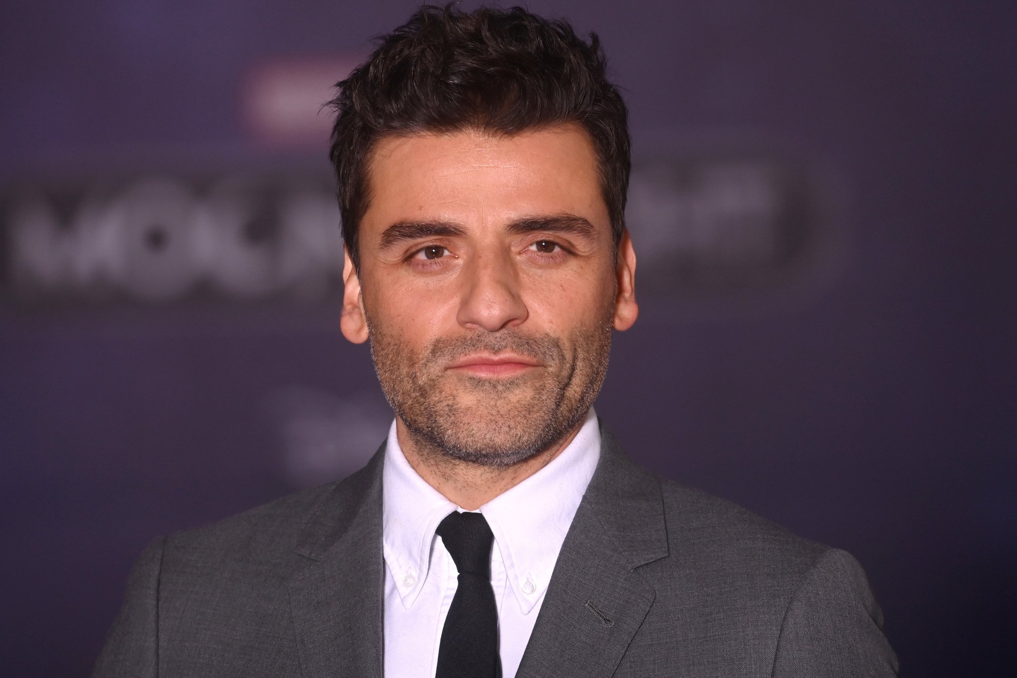 Oscar Isaac at the 'Moon Knight' premiere. He's wearing a white shirt, black tie, and grey jacket. He's looking directly at the camera.