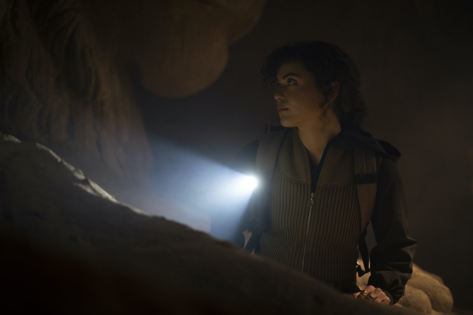 MCU's 'Moon Knight' star May Calamawy, in character as Layla El-Faouly, searches in a dark cave and wears a olive green vest over a black jacket. Layla wears a backpack that has a flashlight on one of the straps.