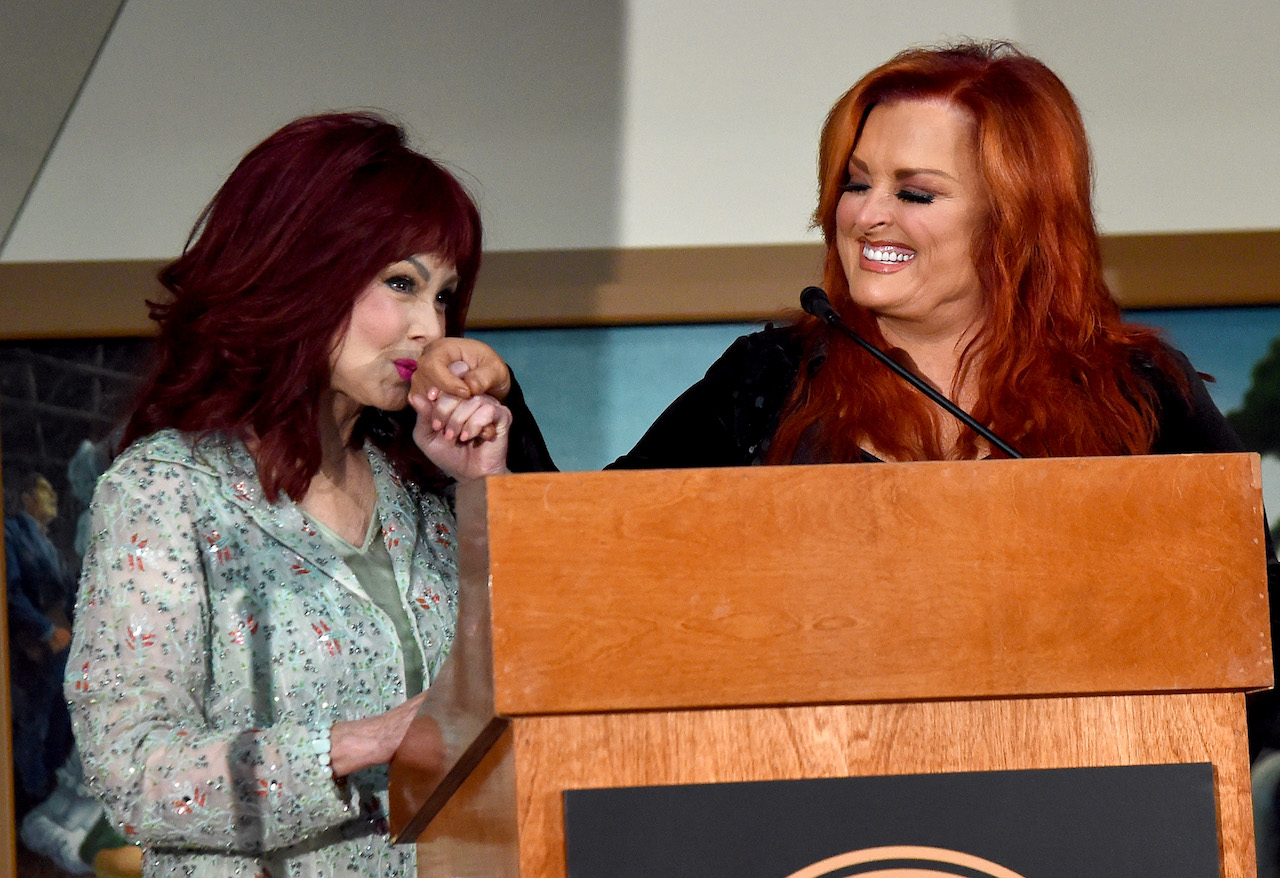 Naomi Judd kisses the hand of her daughter, Wynonna Judd, who is smiling as she watches on