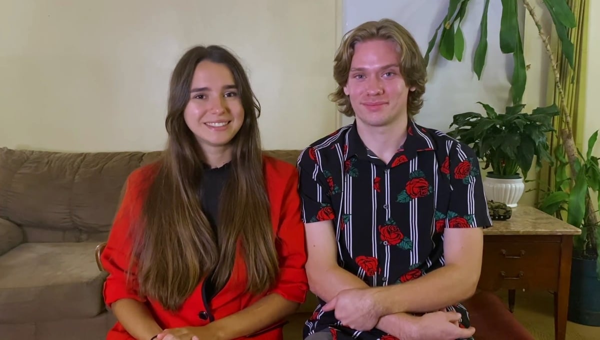 Olga Koshimbetova wearing a red blaser, sitting on a couch with Steven Frend who is wearing a black shirt with red roses on '90 Day Diaries' Season 3. 