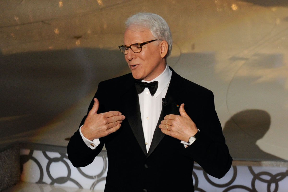 Steve Martin wears a suit and gestures toward himself on stage