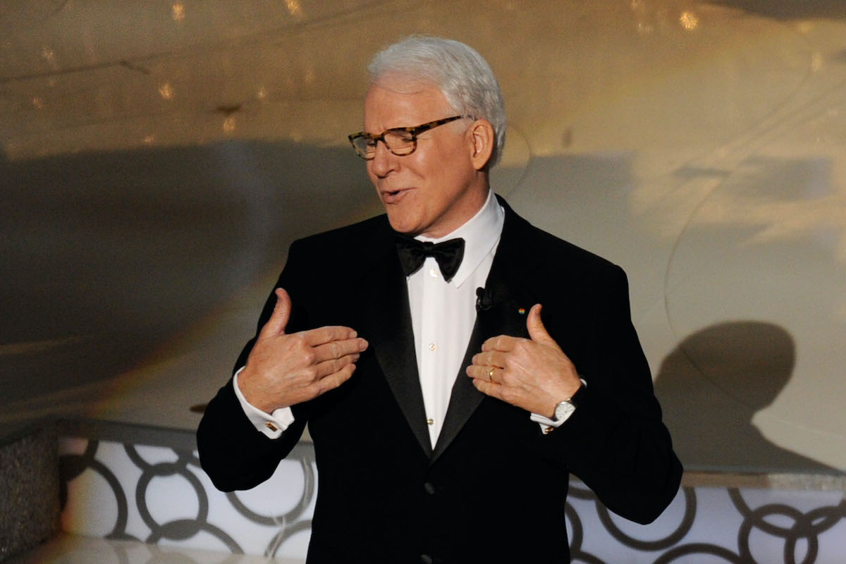 Steve Martin wears a suit and gestures to himself on stage