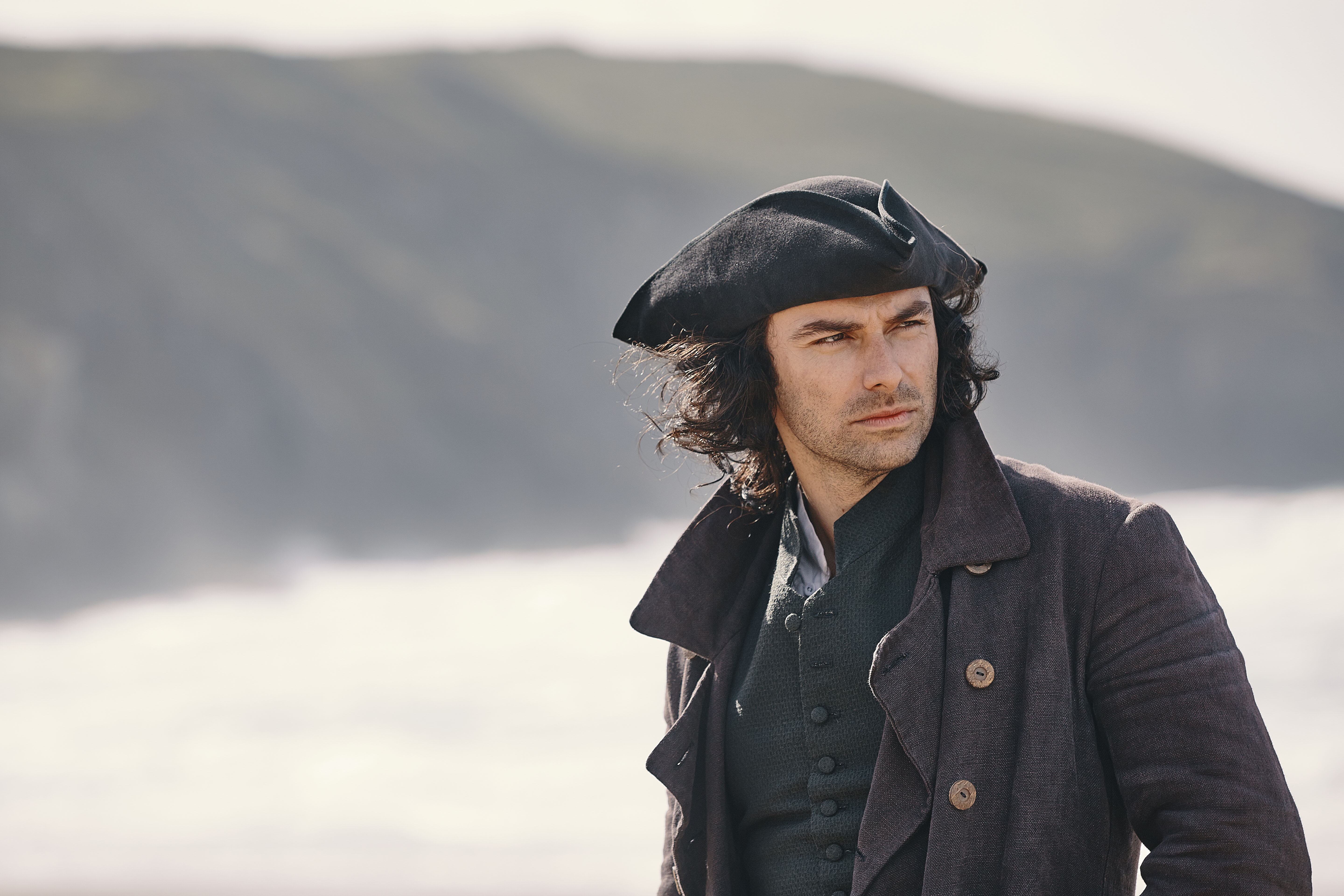 Aidan Turner as Ross Poldark, wearing a tricorn hat and standing in front of a cliff