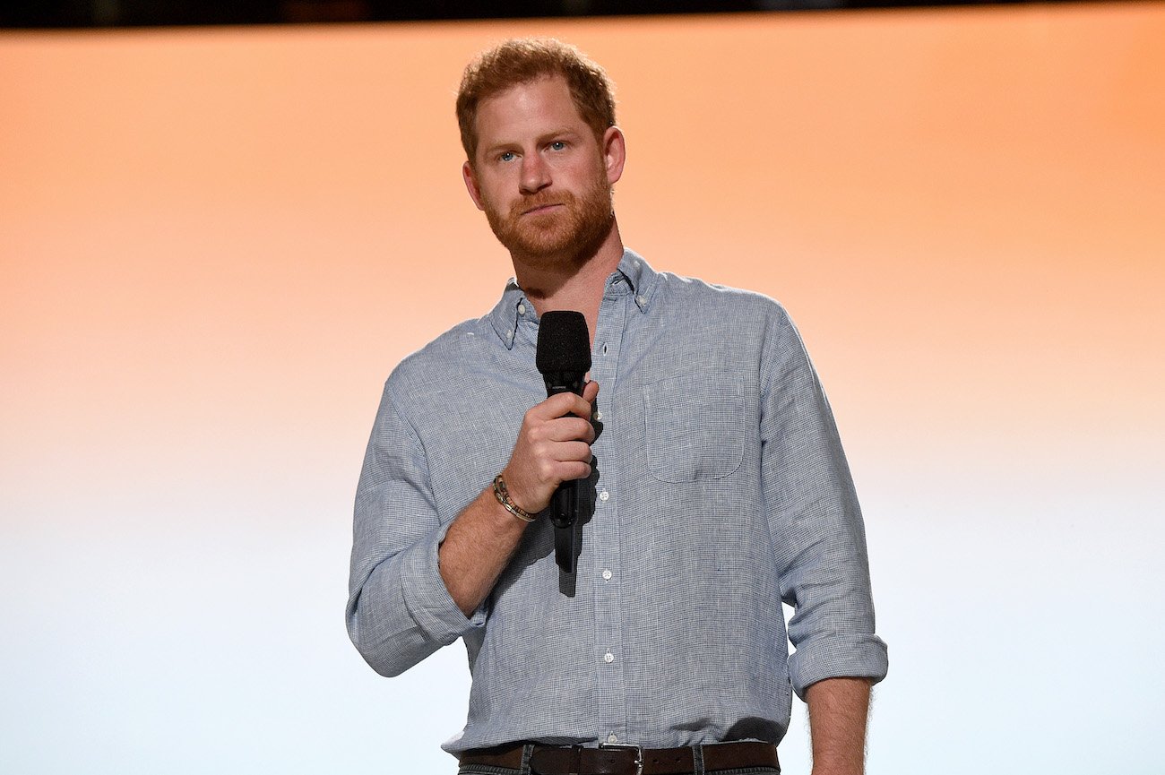 Prince Harry wearing a gray shirt while holding a microphone and standing in front of an orange background