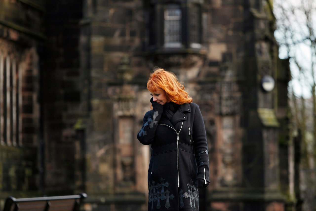 Reba McEntire in a black coat, standing with a hand on her face in an episode of 'Who Do You Think You Are?'