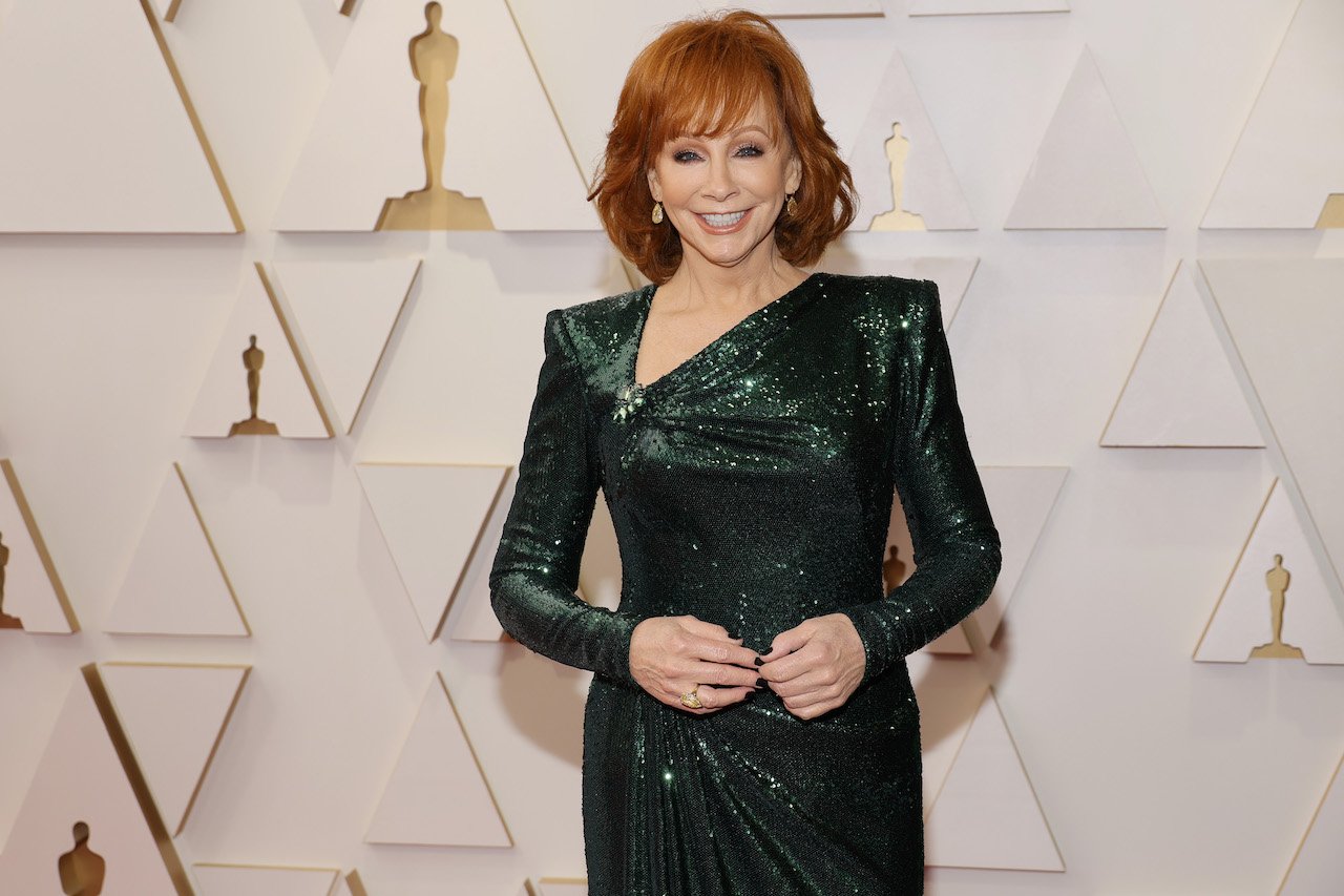 Reba McEntire poses in a dark green dress, smiling in front of an Oscars backdrop