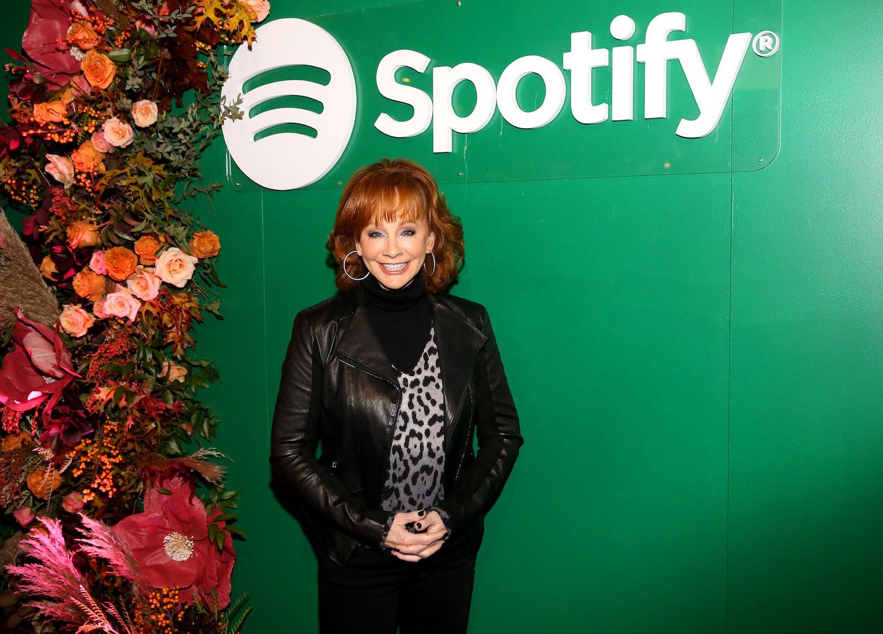 Reba McEntire poses with her hands together in front of her while standing in front of a Spotify backdrop
