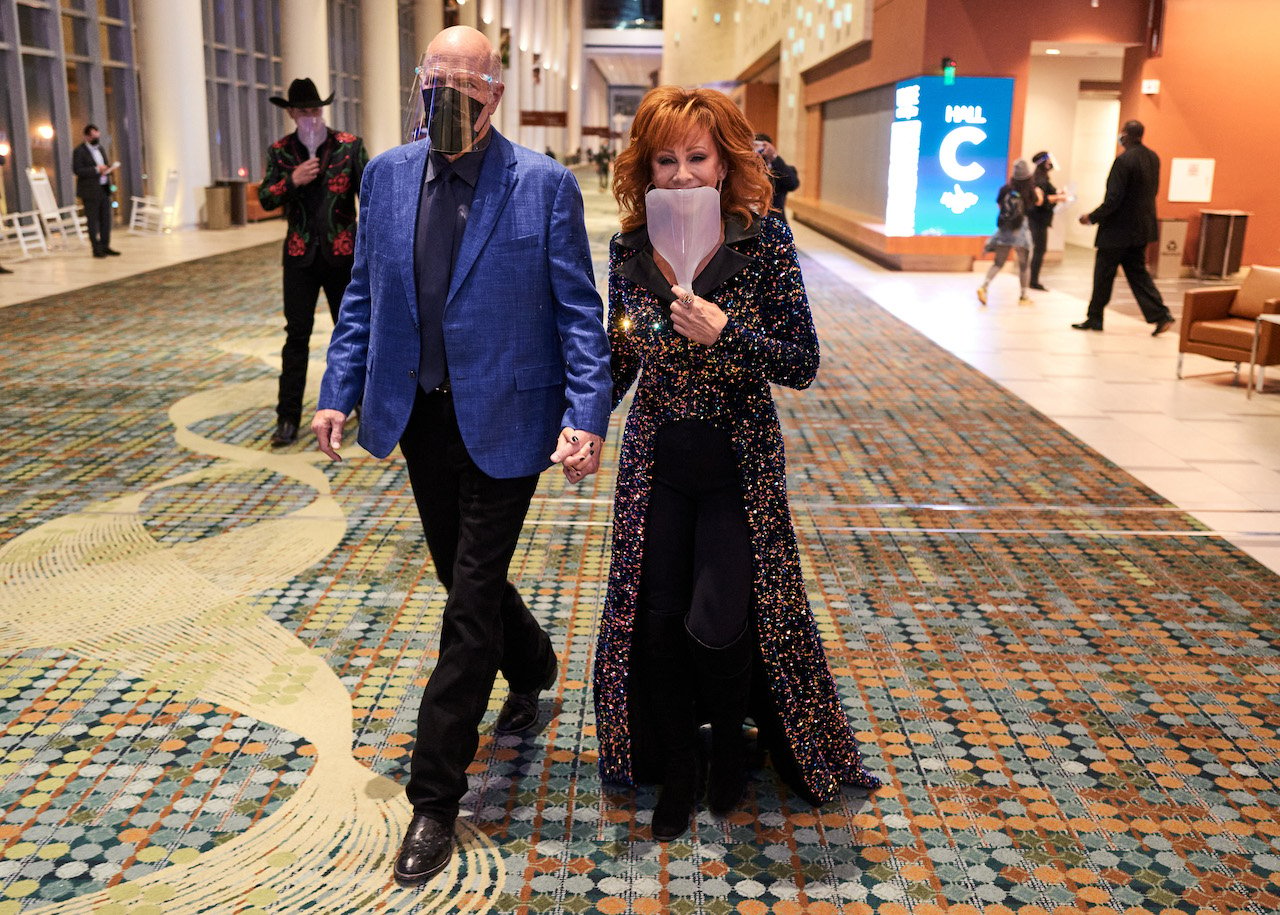 Rex Linn in a blue jacket and a plastic face shiled, holding Reba McEntire's hand as she holds a face shield up to the lower part of her face.