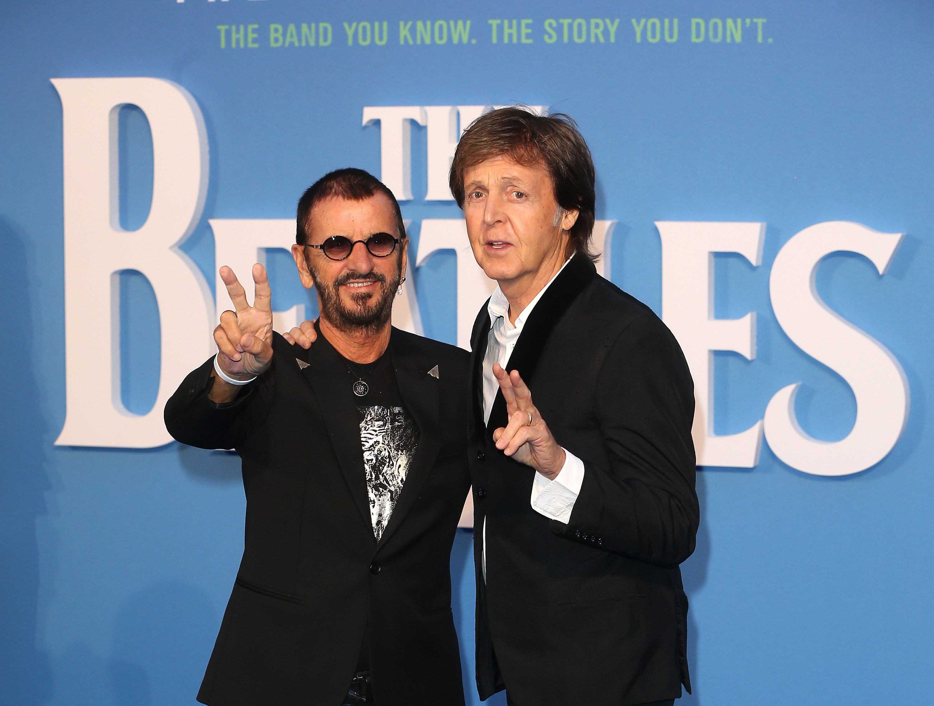 Ringo Starr and Paul McCartney giving peace signs in front of a blue backdrop that says The Beatles in white lettering