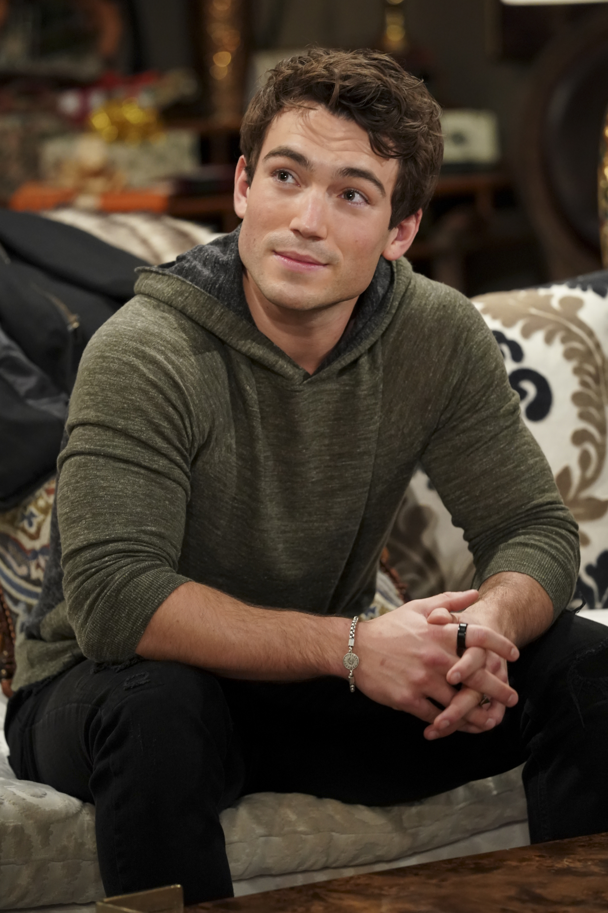 'The Young and the Restless' actor Rory Gibson wearing a grey shirt and black pants sits on a couch.