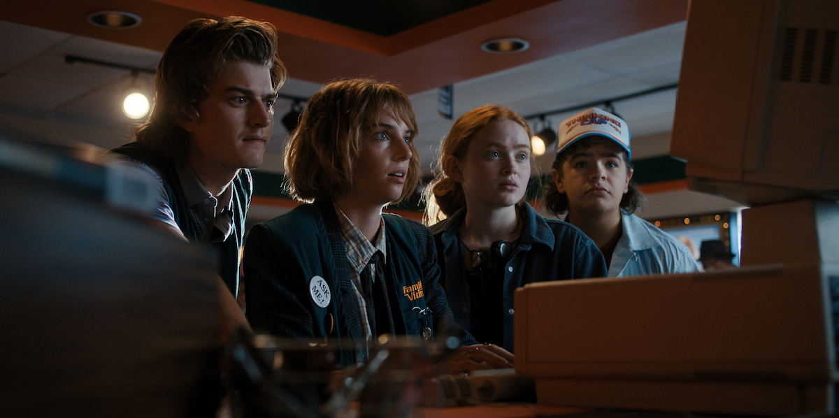 'Stranger Things 4' production still showing Joe Keery, Maya Hawke, Sadie Sink, and Gaten Matarazzo sitting in front of a monitor. 'Stranger Things 4' 80s pop culture moments.