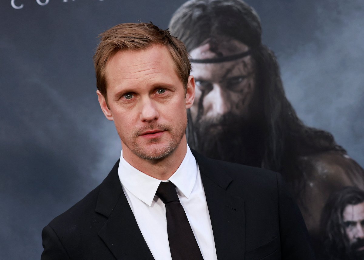 Alexander Skarsgård wears a black suit and poses in front of an image of himself in ‘The Northman’